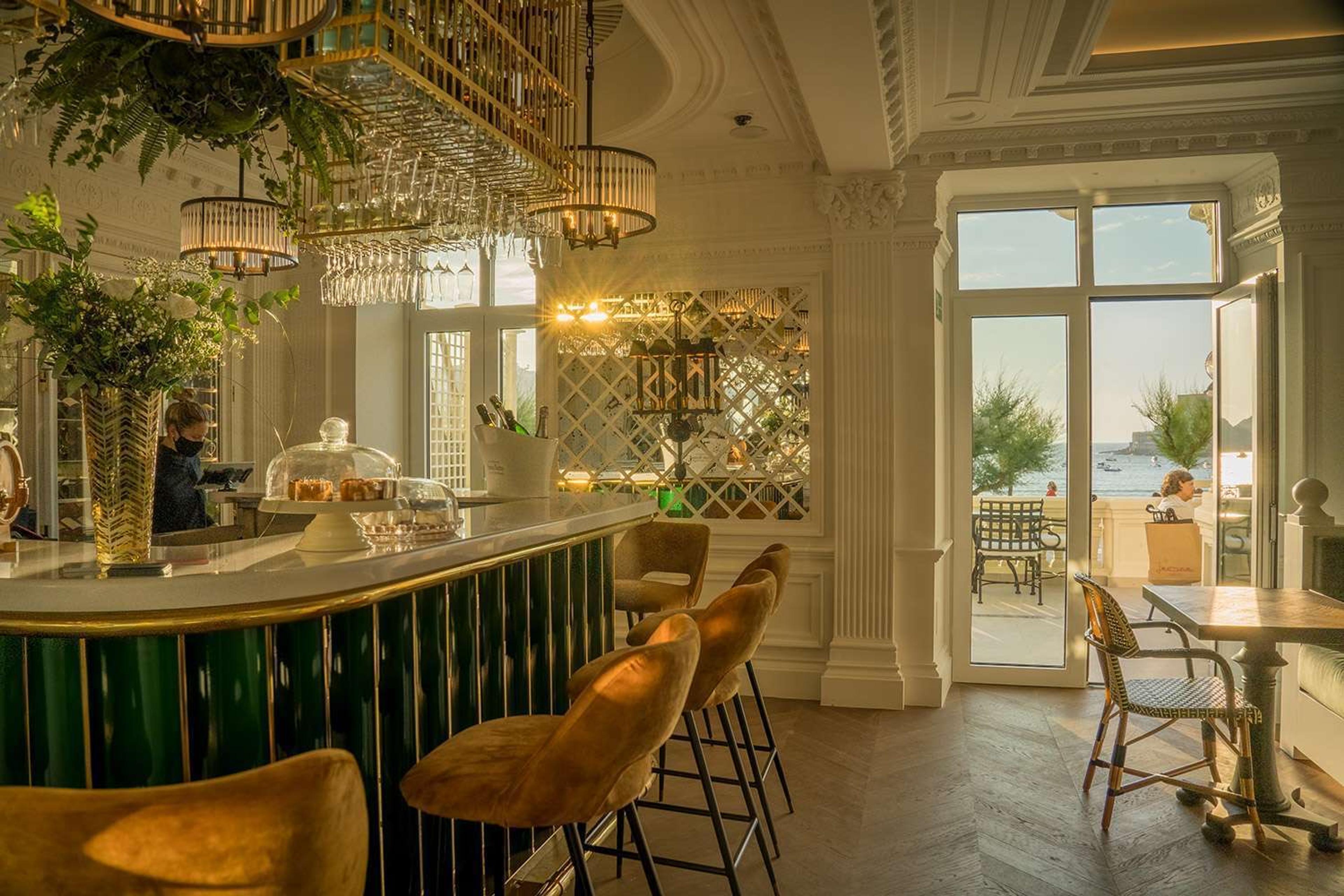 A hotel bar with green and brass panelling, velvet stools, hanging glassware, cakes under glass domes, and open doors to a sunlit terrace.