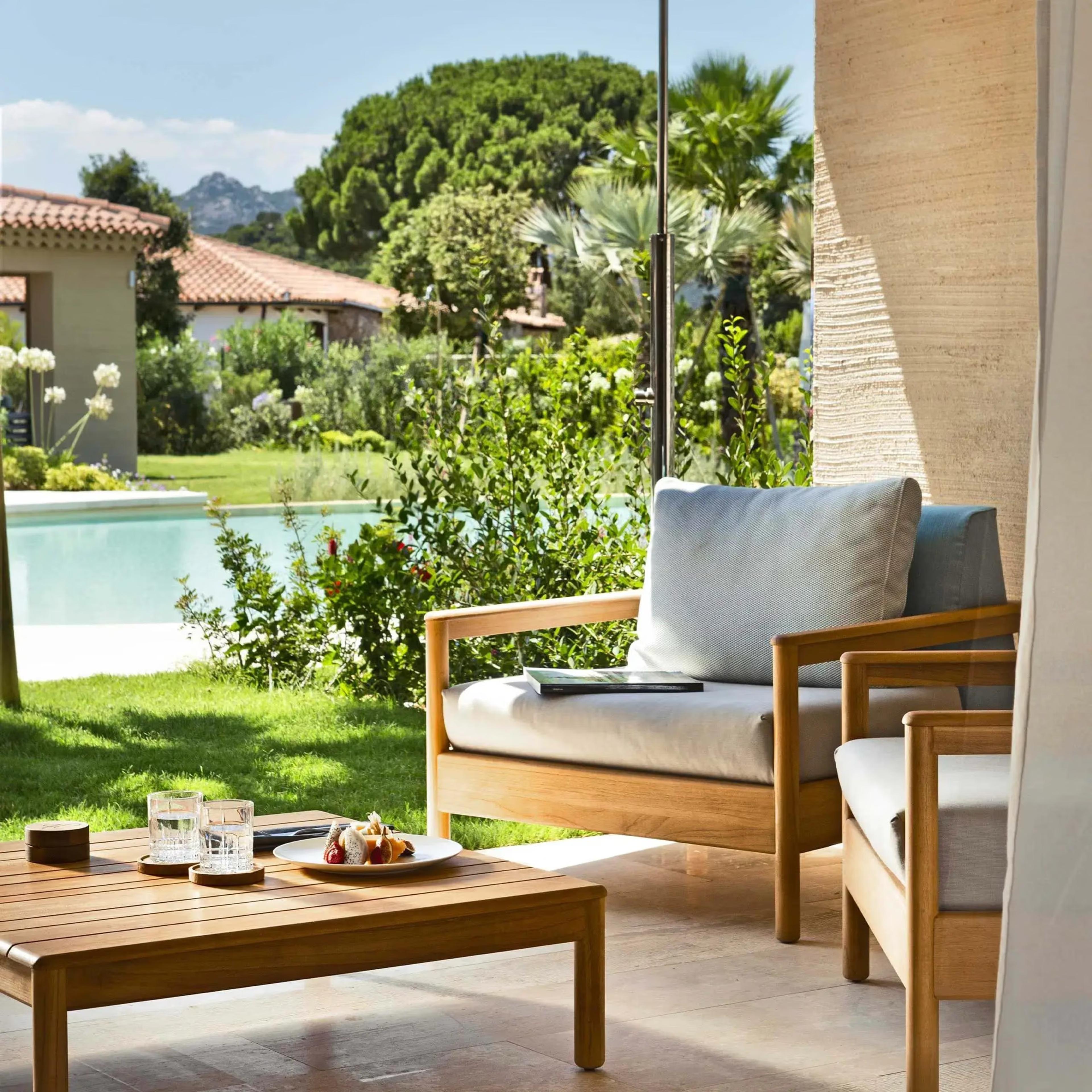Wicker lounge chairs and a coffee table with breakfast on a terrace beside a swimming pool, garden and mountains beyond.