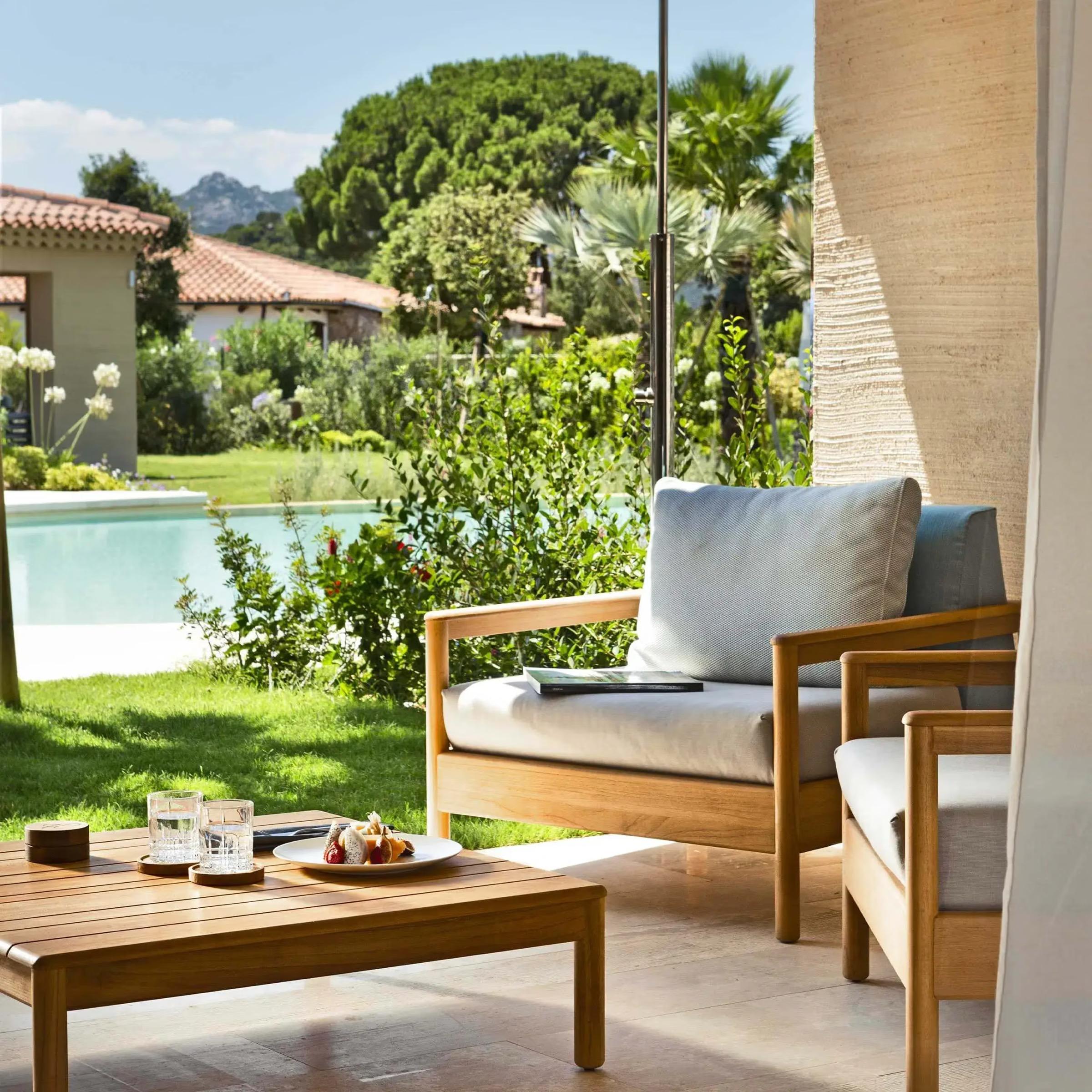 Wicker lounge chairs and a coffee table with breakfast on a terrace beside a swimming pool, garden and mountains beyond.