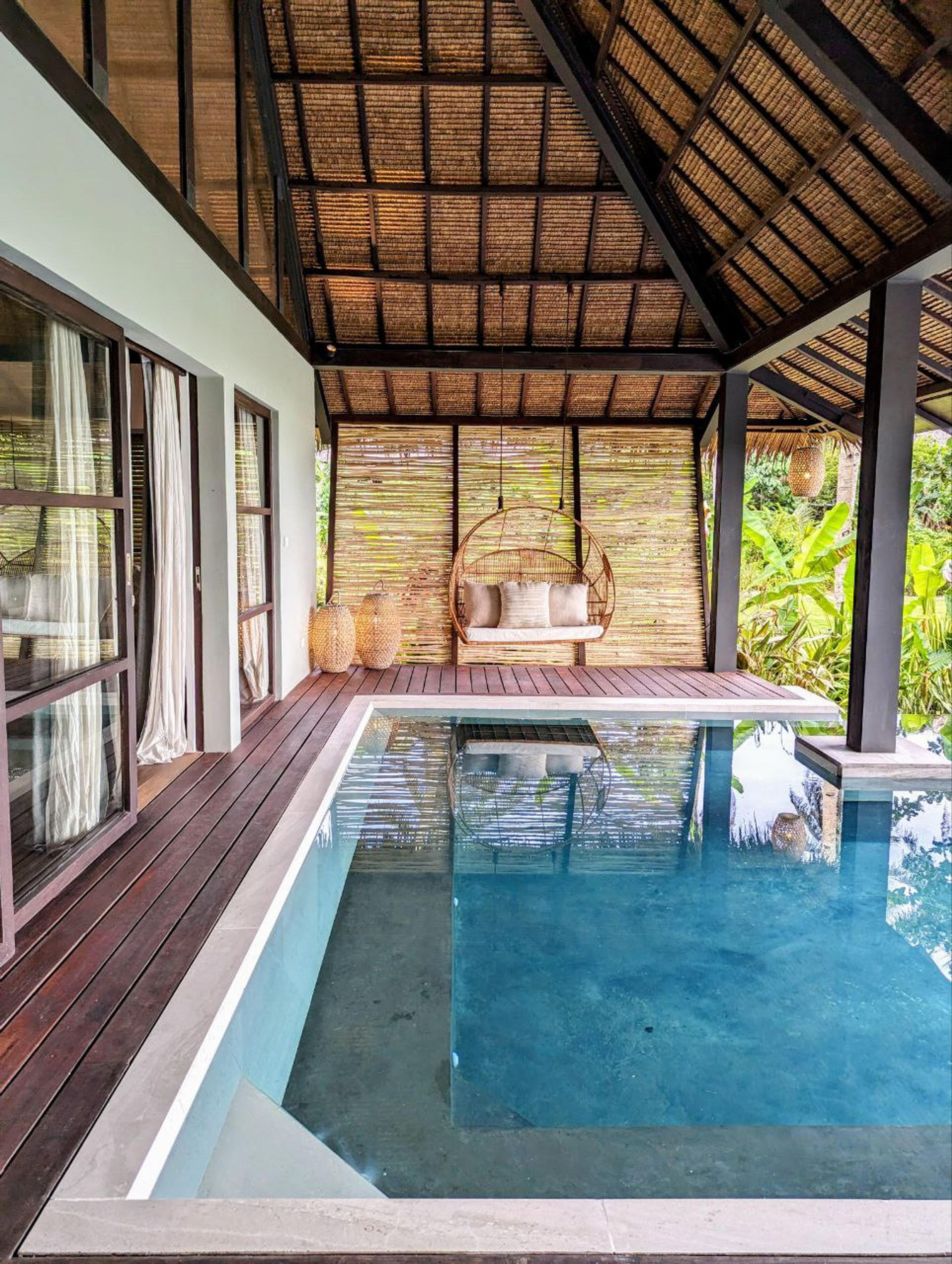 A villa with a thatched roof stands beside its own pool, a hanging egg chair on the deck and plants crowding the edge.