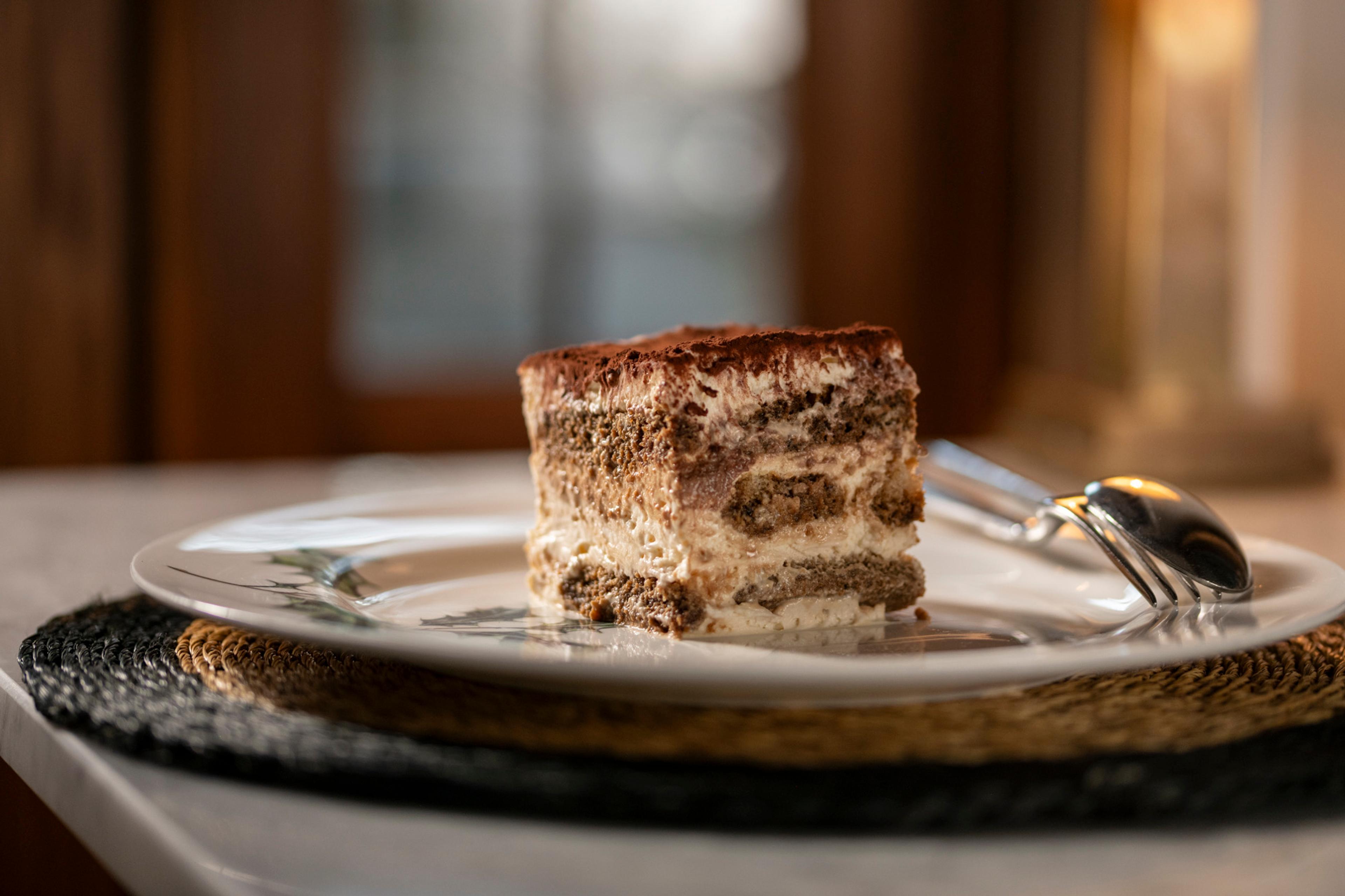 A square of tiramisu on a white plate with a fork and spoon, on a table with woven placemats.