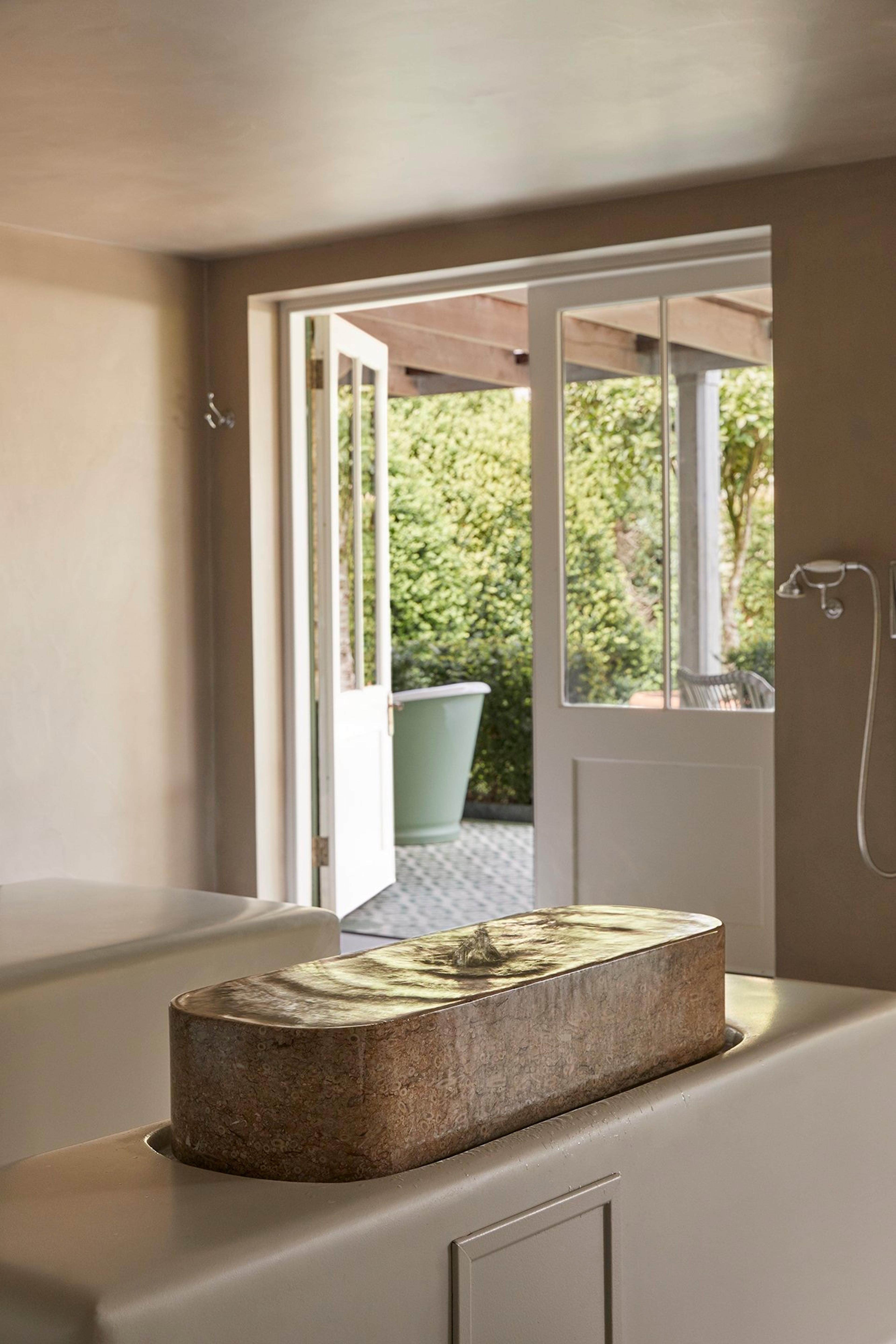 A rectangular stone bath built into a ledge with water rippling across it, beside open doors leading out to a planted courtyard.