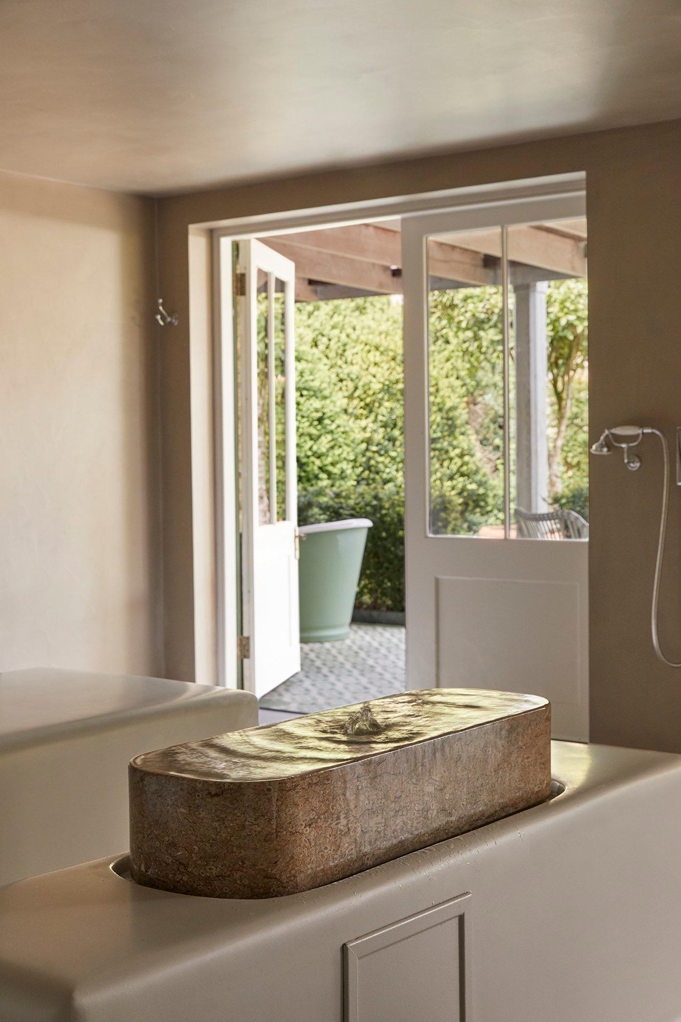 A rectangular stone bath built into a ledge with water rippling across it, beside open doors leading out to a planted courtyard.