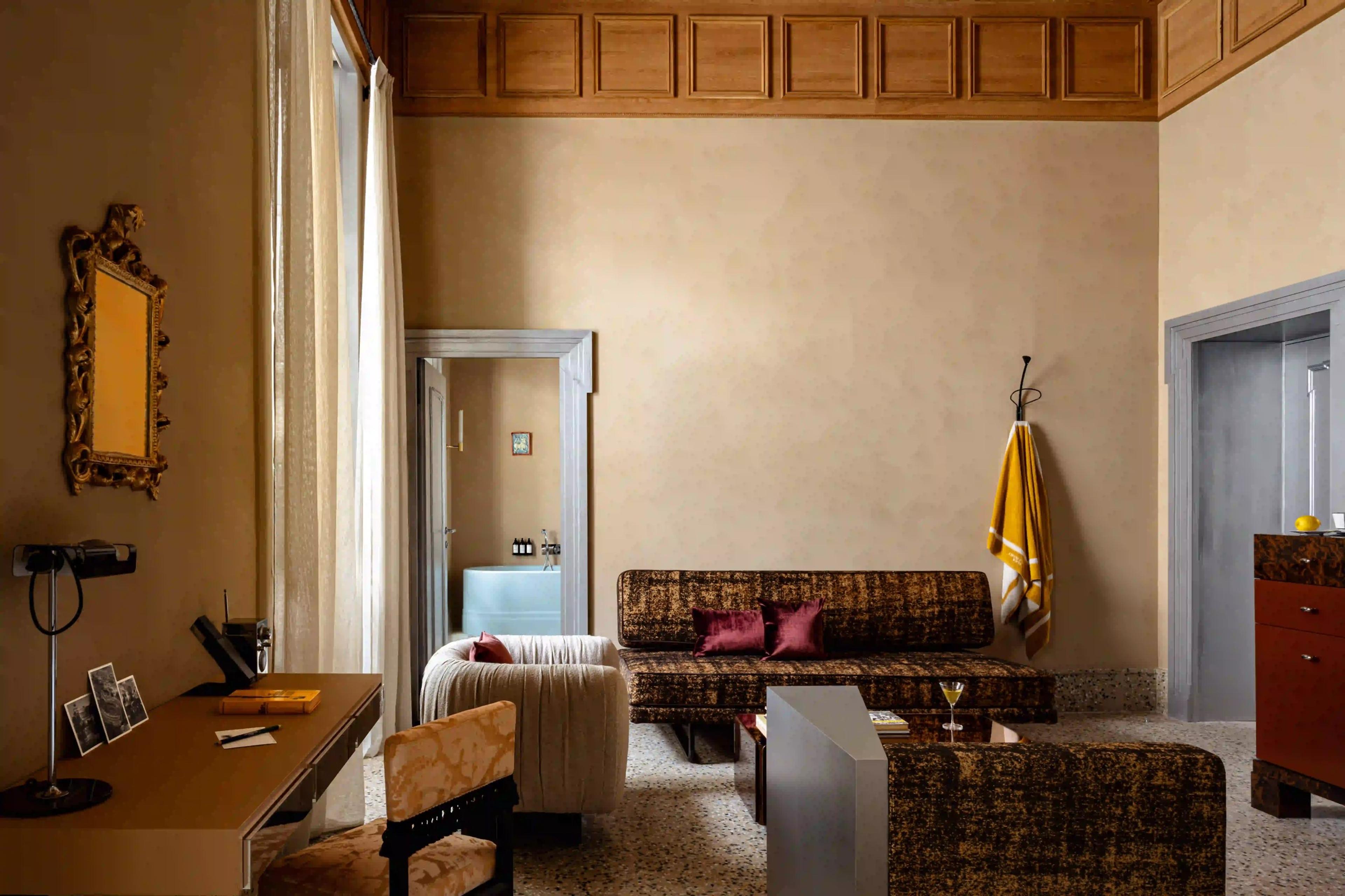 A high-ceilinged sitting room with a dark patterned sofa, a chaise armchair, a desk with photographs and a lamp, and a pale blue bathtub visible through an open doorway.