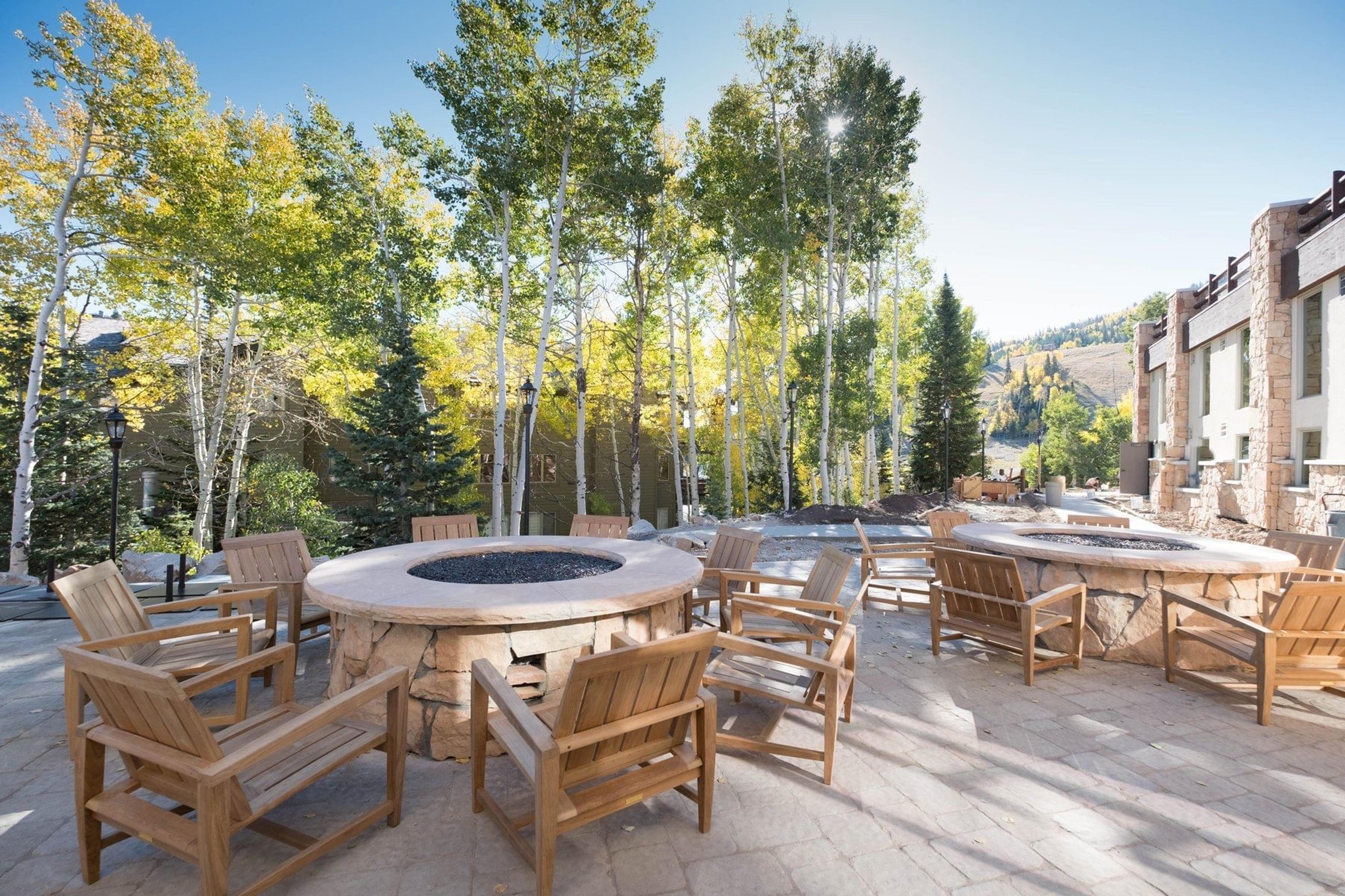 two round stone-edged fire pits surrounded by wooden chairs on a stone patio, set among white-barked aspens in daylight