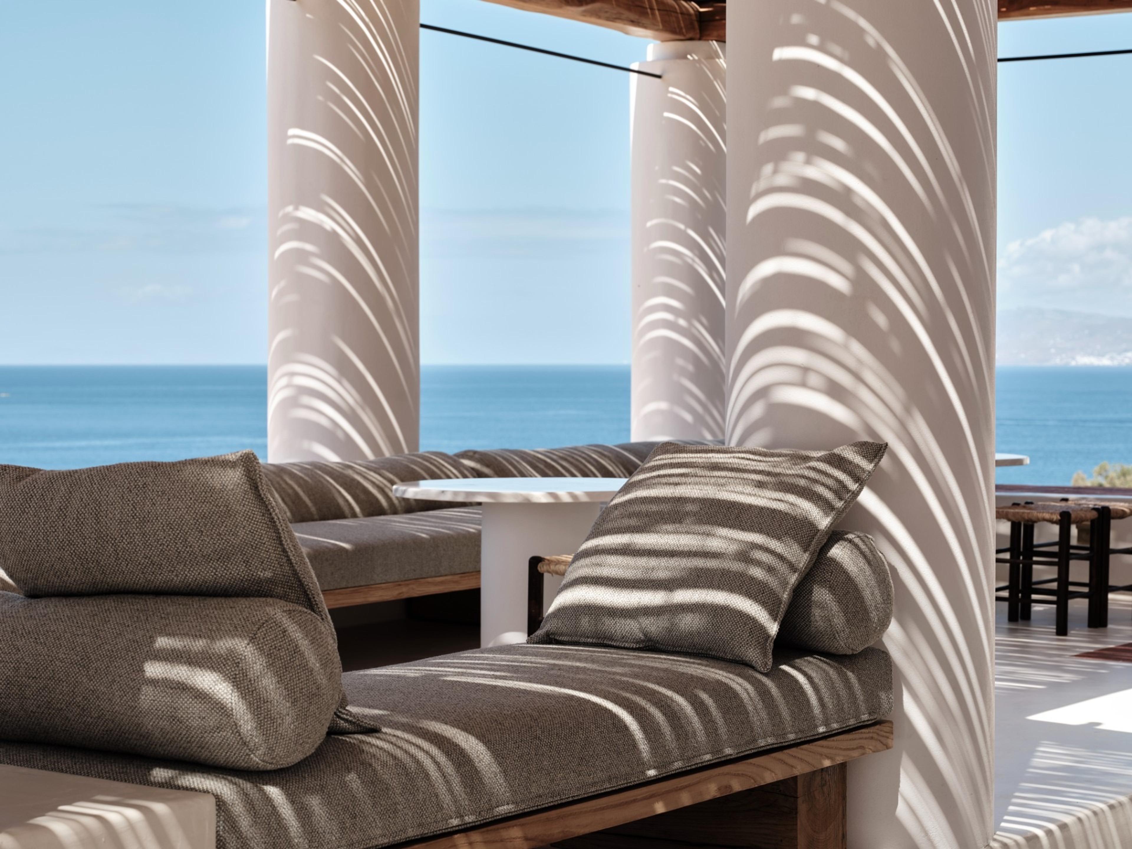 A row of cushioned daybeds sits under a shaded pergola with round white columns, striped in sunlight, looking out over the sea.