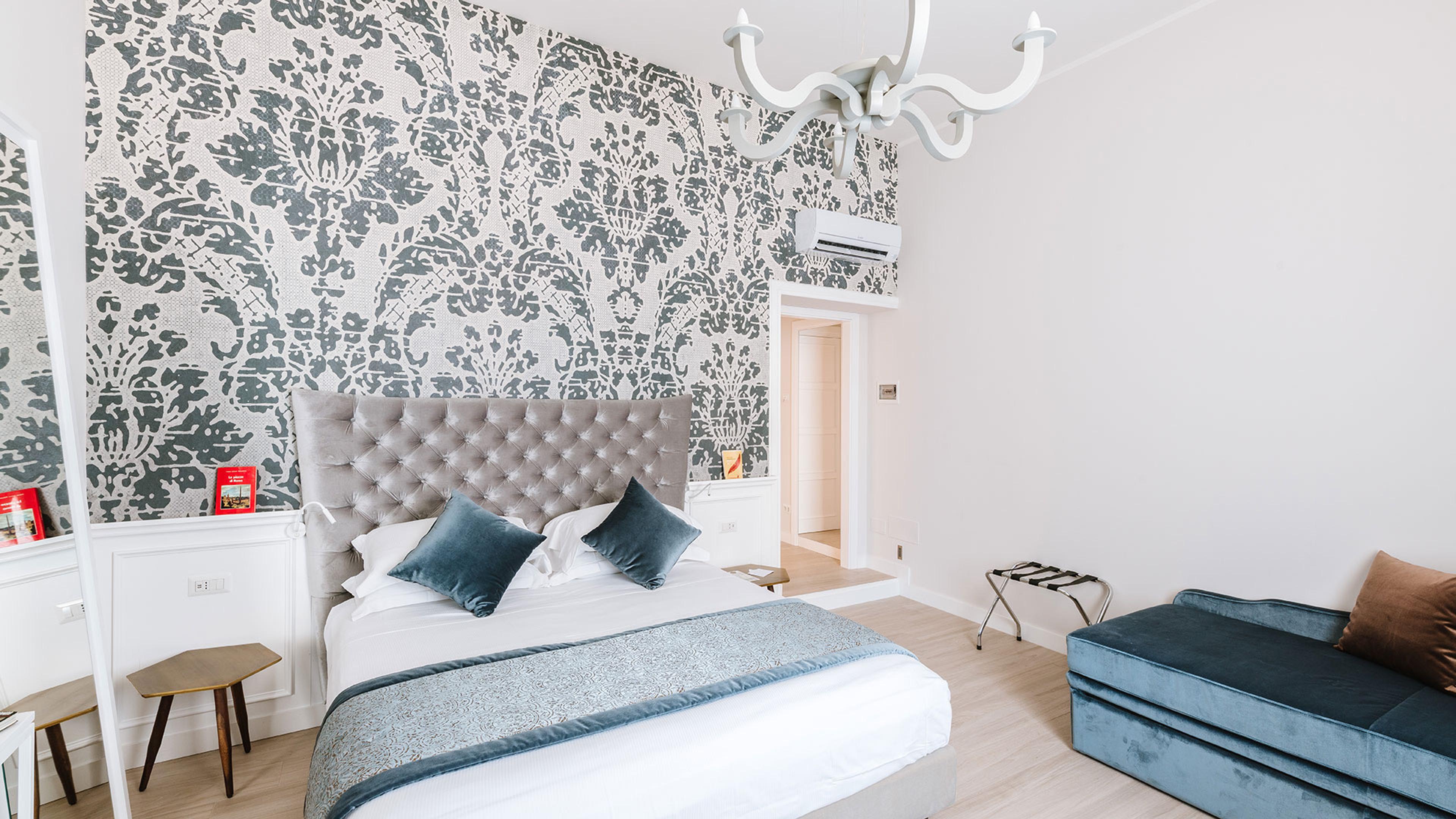 A bedroom with grey damask wallpaper, a tufted grey headboard, and a dark blue chaise sofa against the opposite wall.