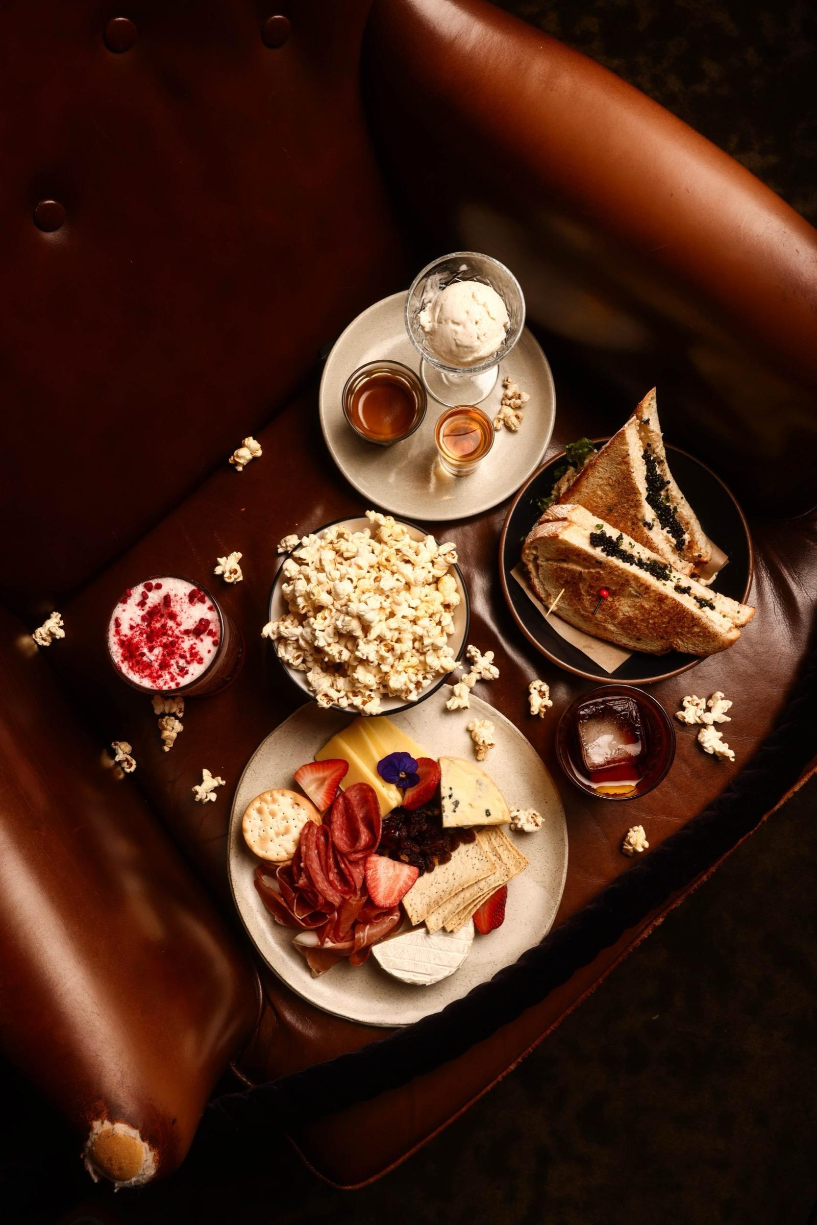 A flat lay on a leather banquette of popcorn, a charcuterie and cheese plate, a toasted sandwich, affogato, and two cocktails.