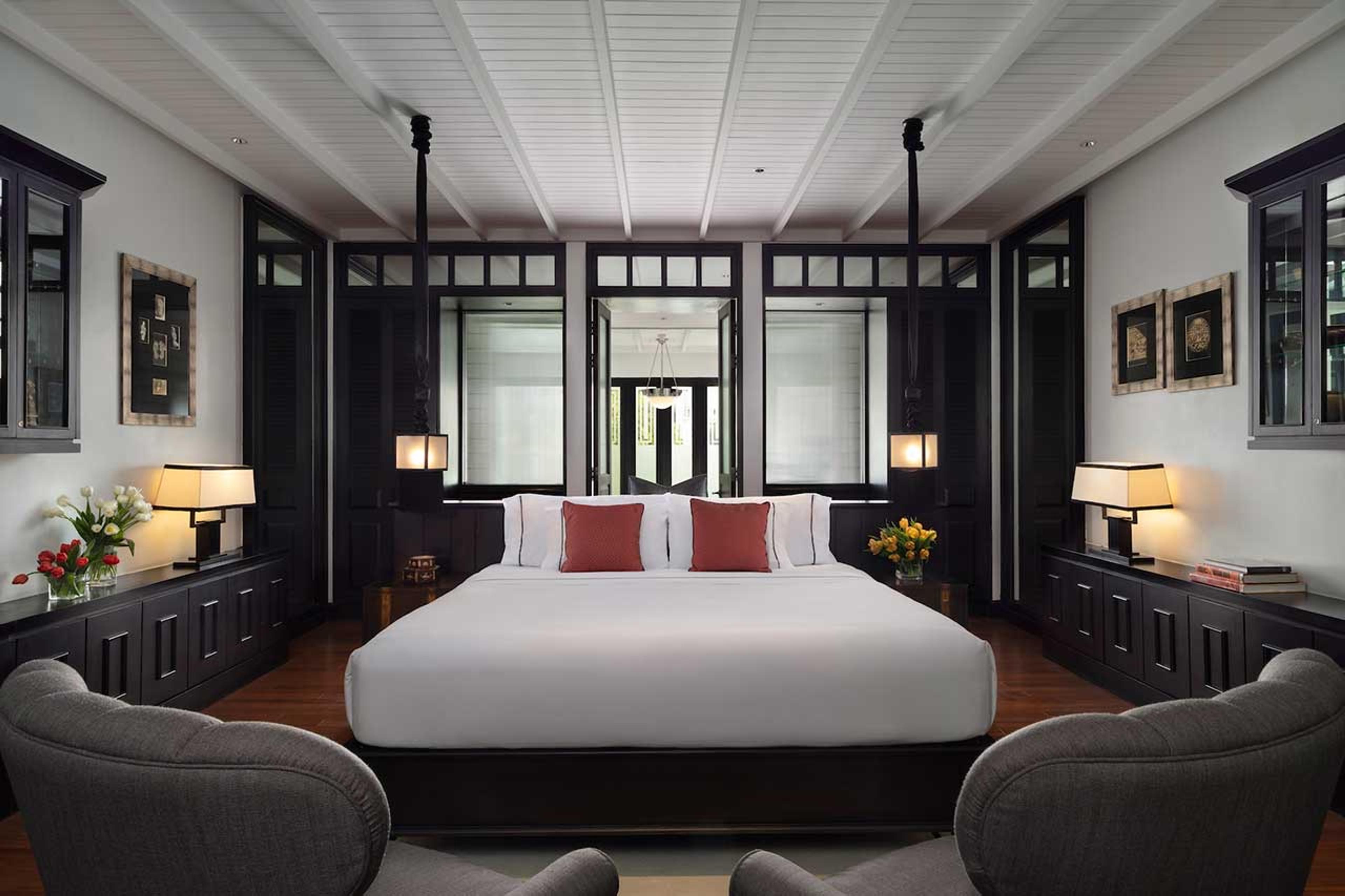 A wide bed with white linen and red cushions sits between two grey armchairs, flanked by dark wood cabinets, mirrored black-framed doors and hanging lanterns.