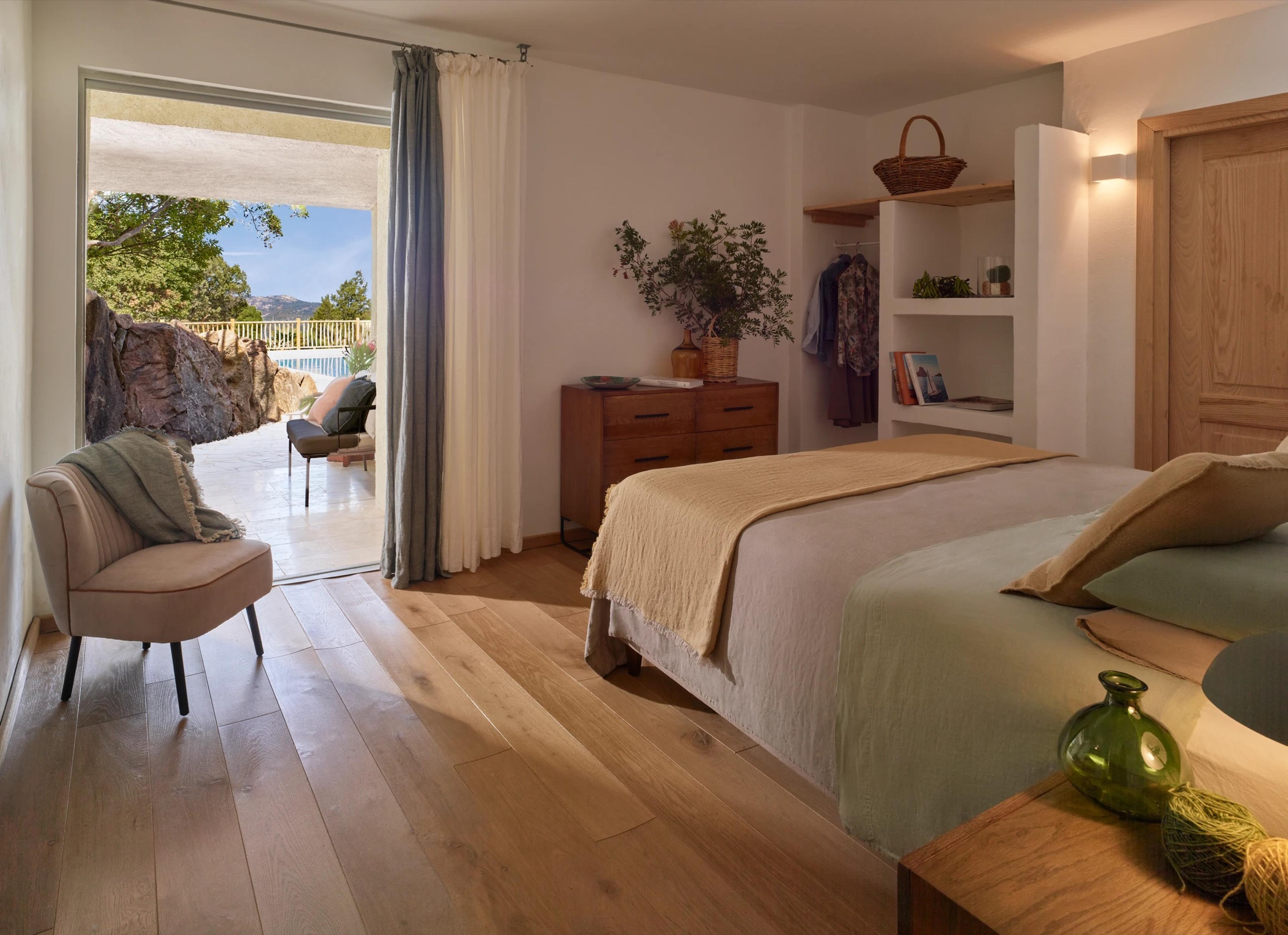 a bedroom with a woven armchair, a wooden dresser and a bed, the sliding door open to a stone terrace and a pool beyond