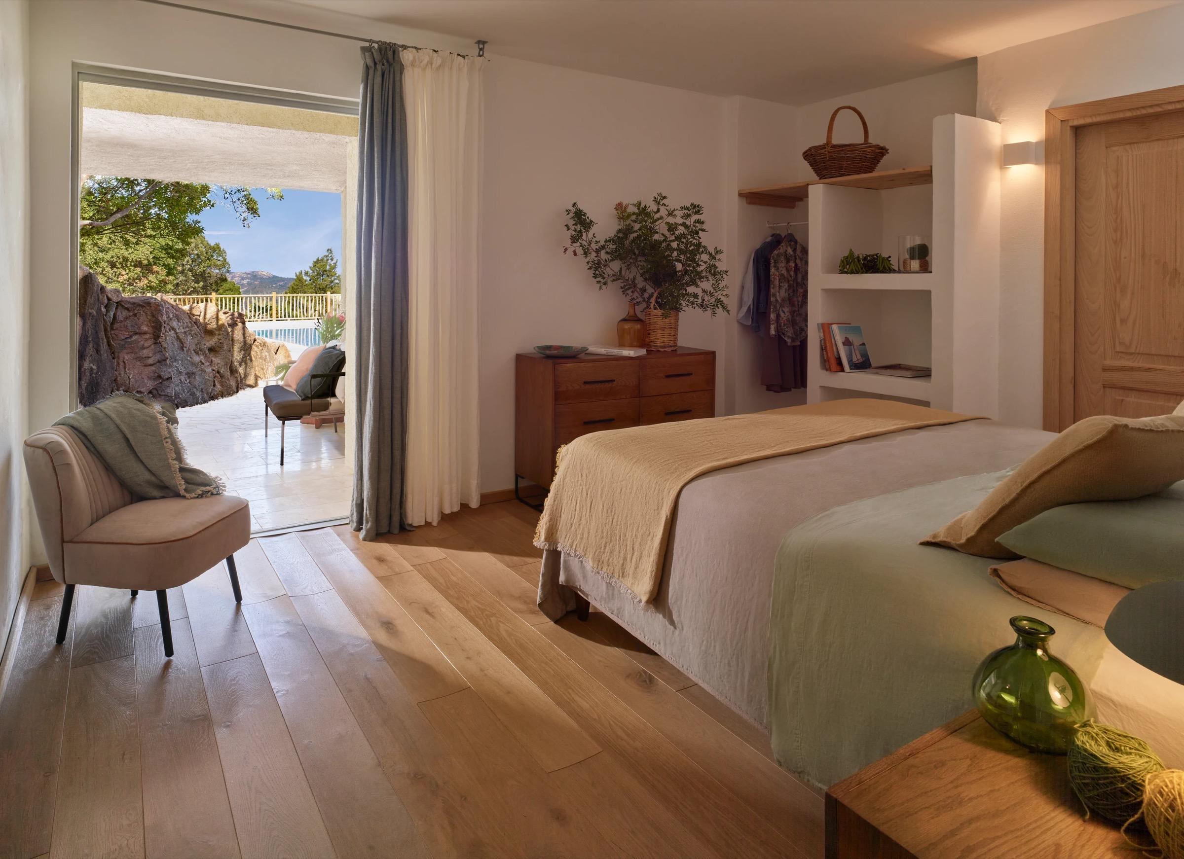a bedroom with a woven armchair, a wooden dresser and a bed, the sliding door open to a stone terrace and a pool beyond
