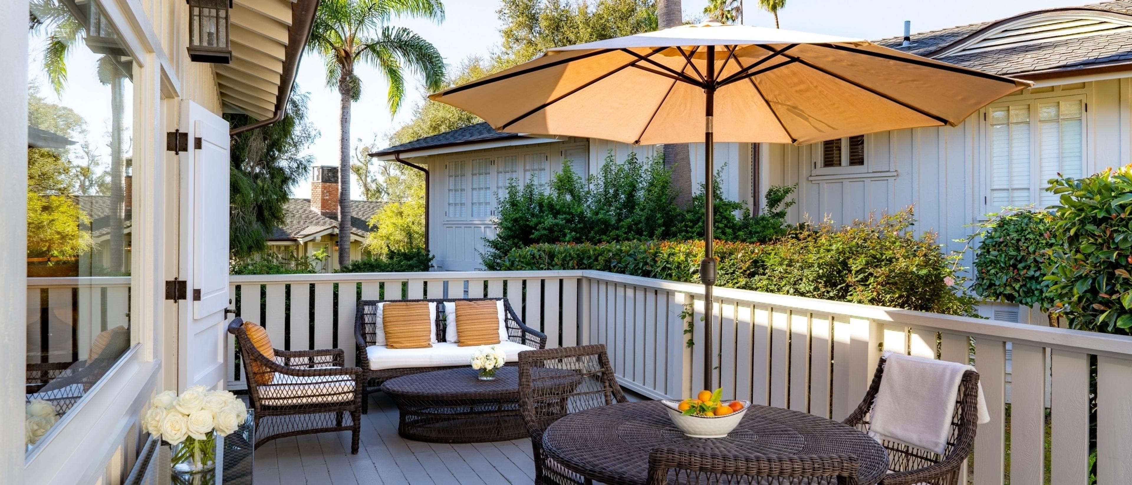 A furnished outdoor terrace with a sofa, armchairs and a shaded dining table holding a bowl of citrus, framed by a white balustrade and a neighbouring tiled-roof building.