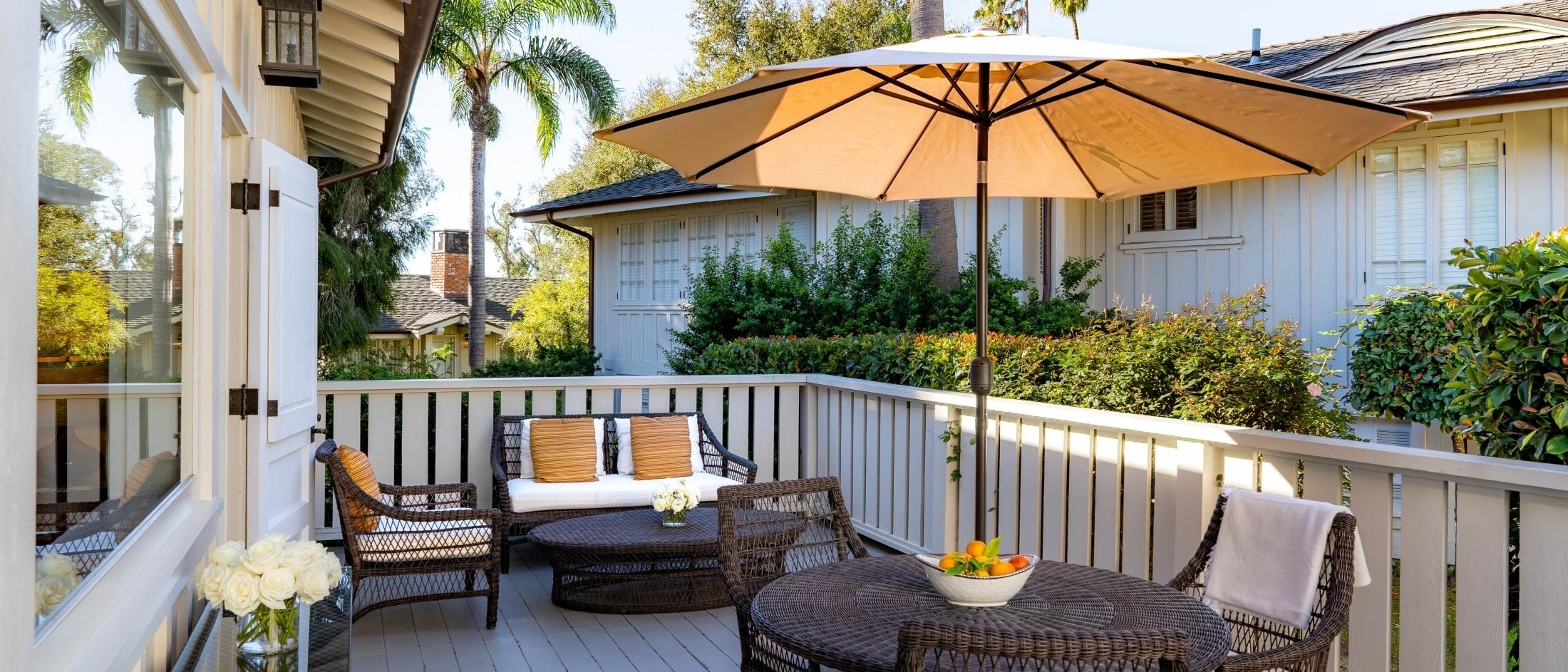 A furnished outdoor terrace with a sofa, armchairs and a shaded dining table holding a bowl of citrus, framed by a white balustrade and a neighbouring tiled-roof building.