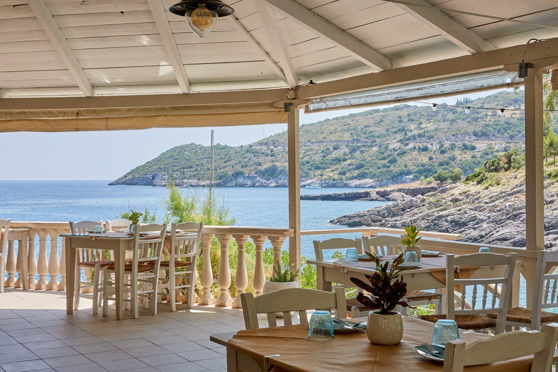 An open taverna terrace of white wooden tables under a timber roof, looking out over a blue cove and wooded headland
