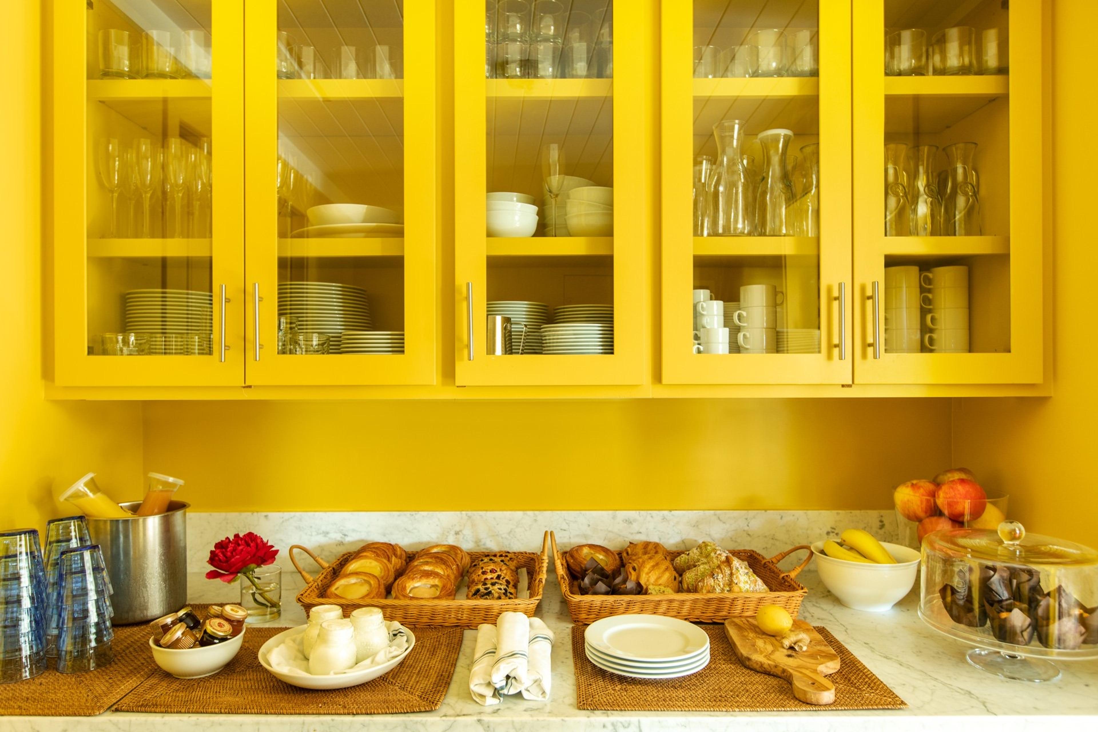 A marble counter beneath yellow glass-fronted cabinets, set with baskets of pastries, a bowl of fruit, a glass-domed cake stand of muffins, and glasses of juice.