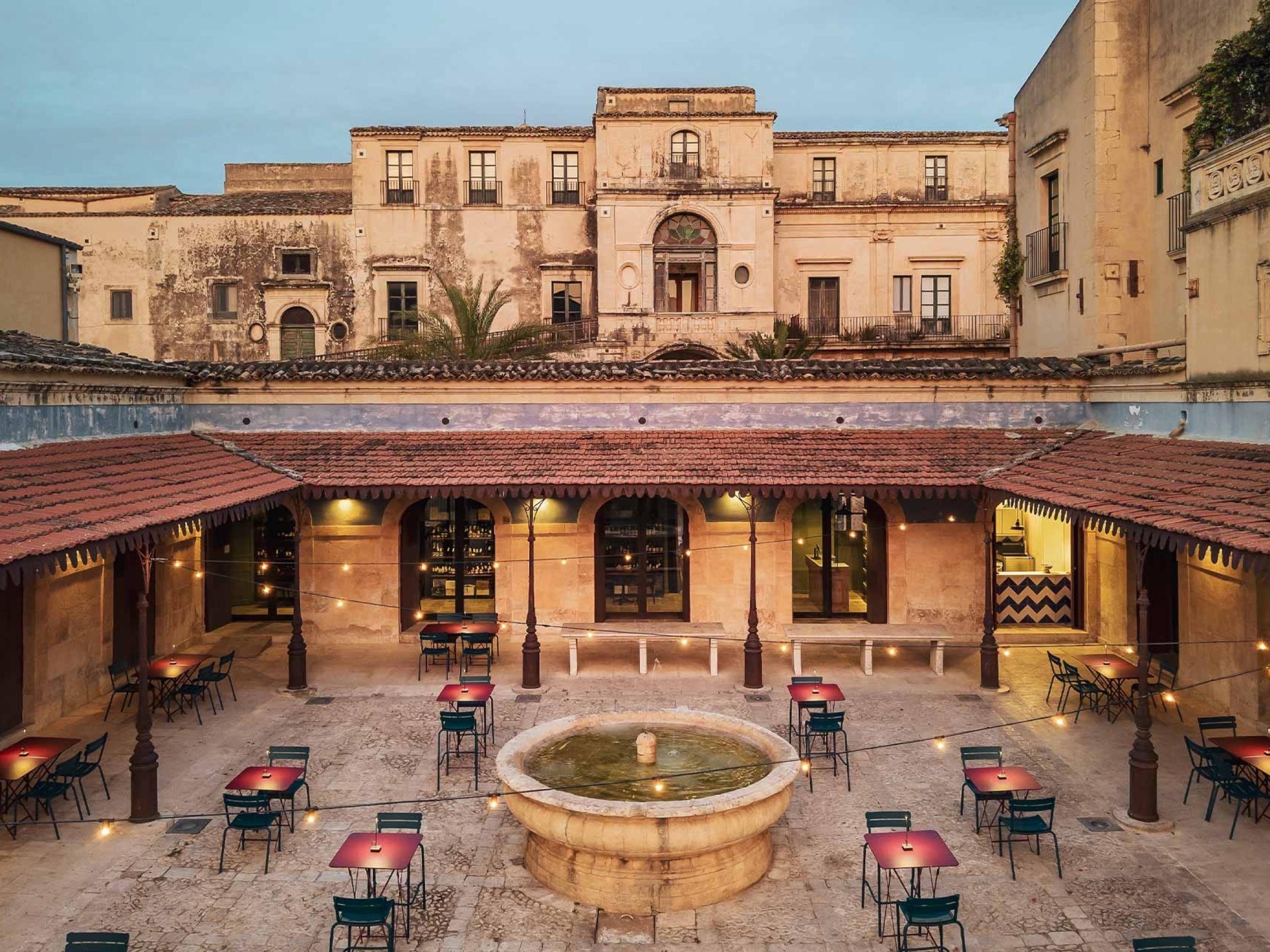 The courtyard of Palazzo Nicolaci in Noto at dusk, café tables round a stone fountain under the Baroque palace front, at Seven Rooms Villadorata