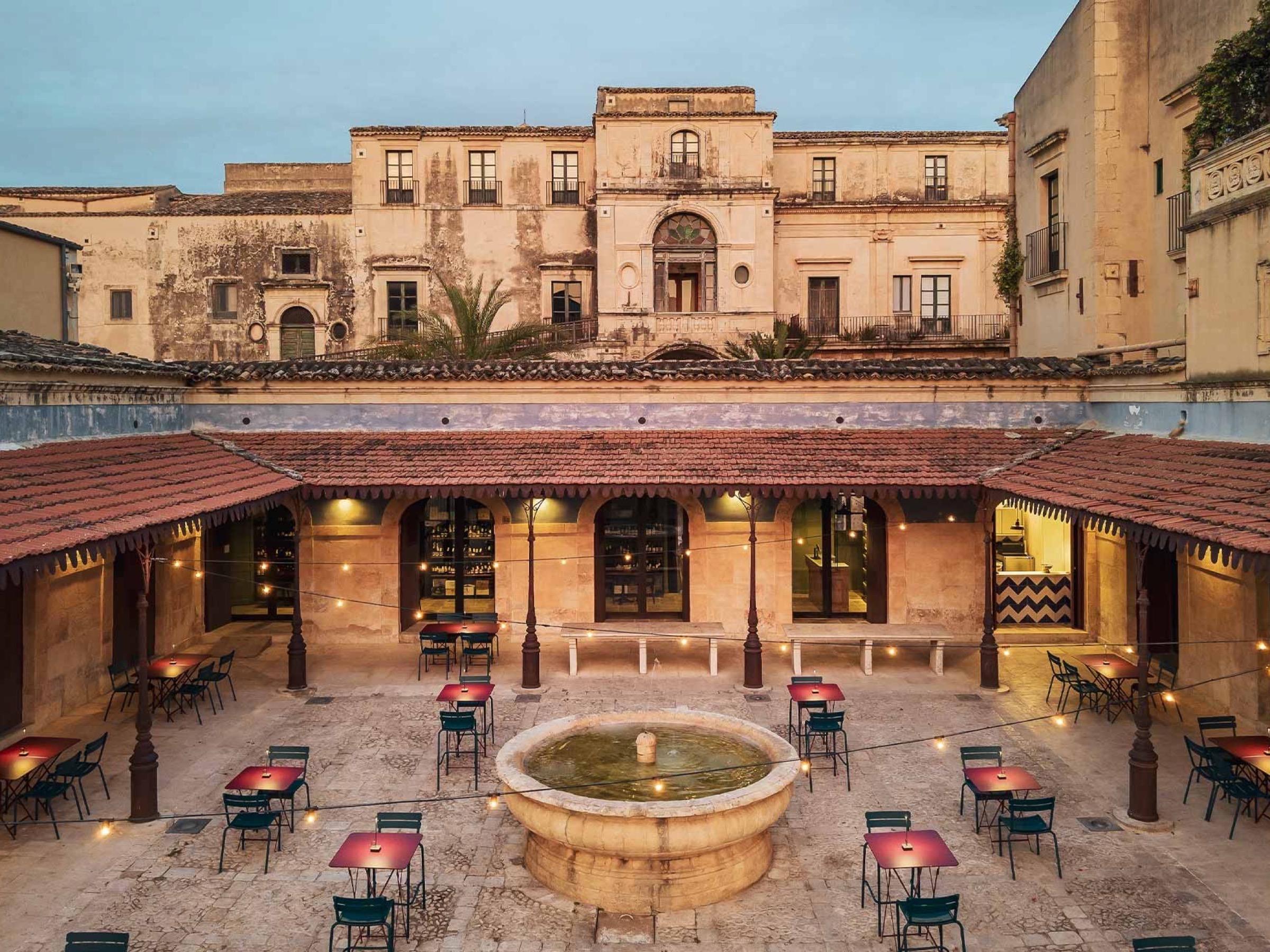 The courtyard of Palazzo Nicolaci in Noto at dusk, café tables round a stone fountain under the Baroque palace front, at Seven Rooms Villadorata