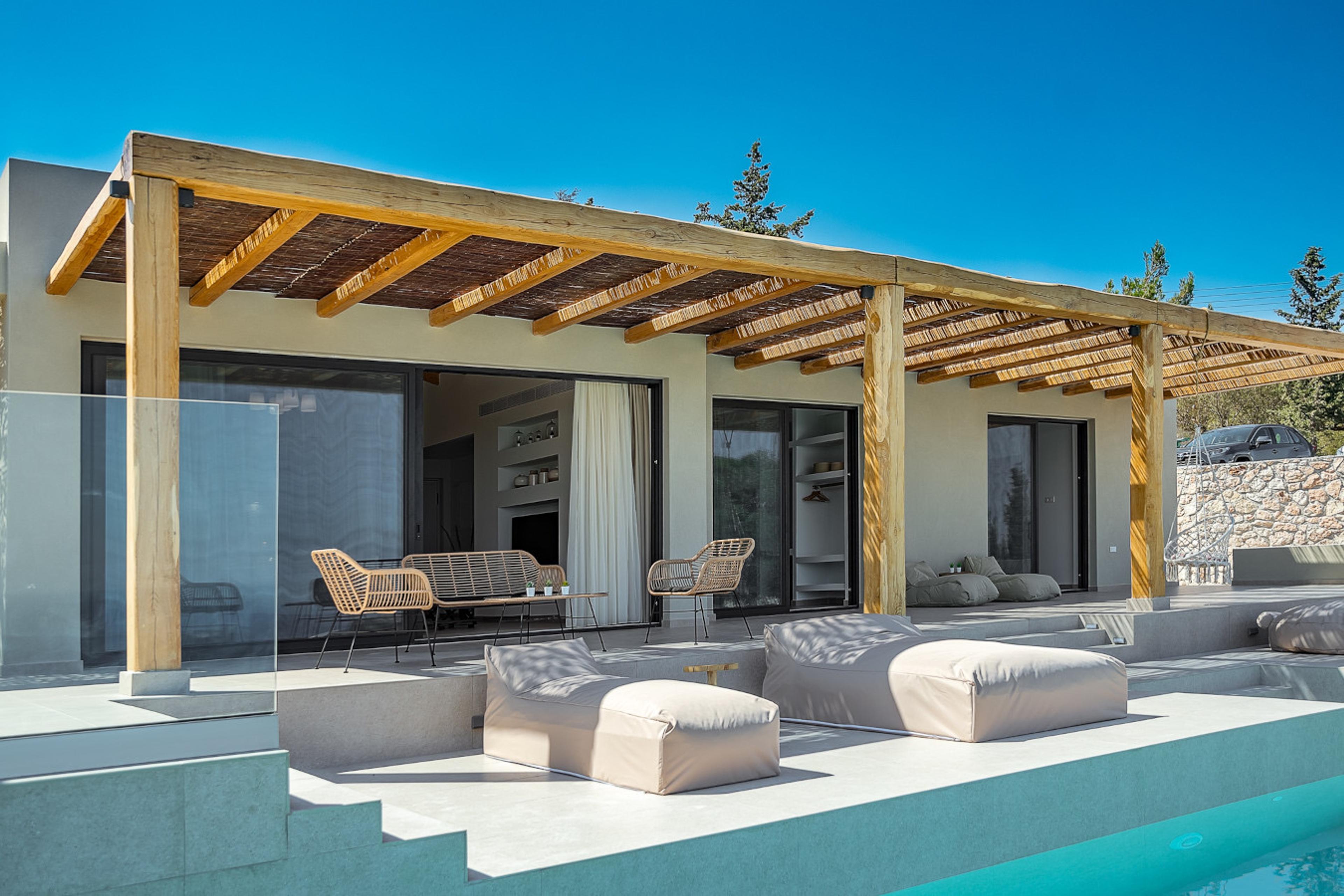 A single-storey villa with a wooden pergola roof along its terrace, a glass-fenced plunge pool, pale cushioned loungers and a car parked beyond a low stone wall.