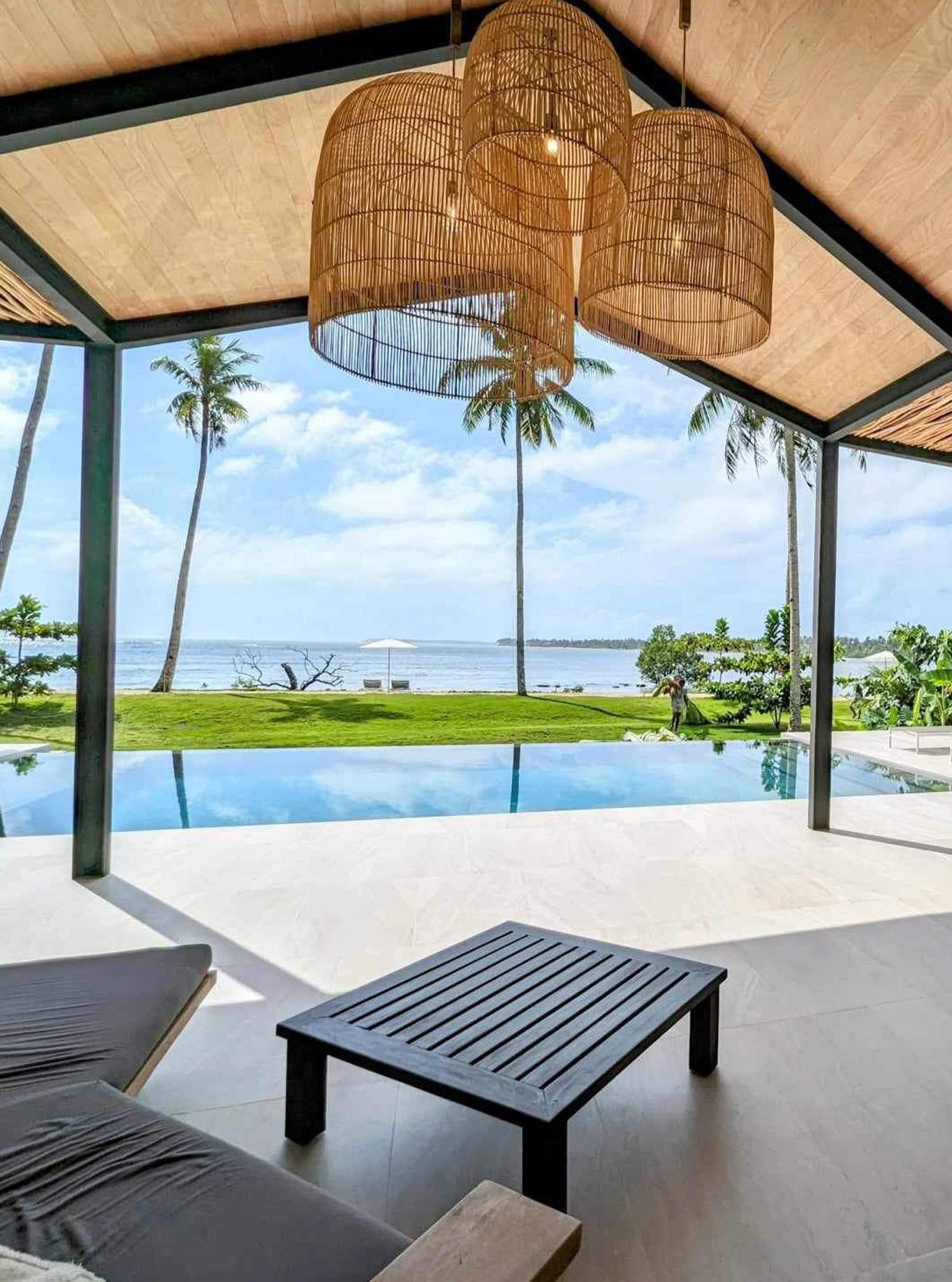 A covered terrace frames a view of the sea past a lawn and palm trees, with sun loungers and a woven lantern overhead.