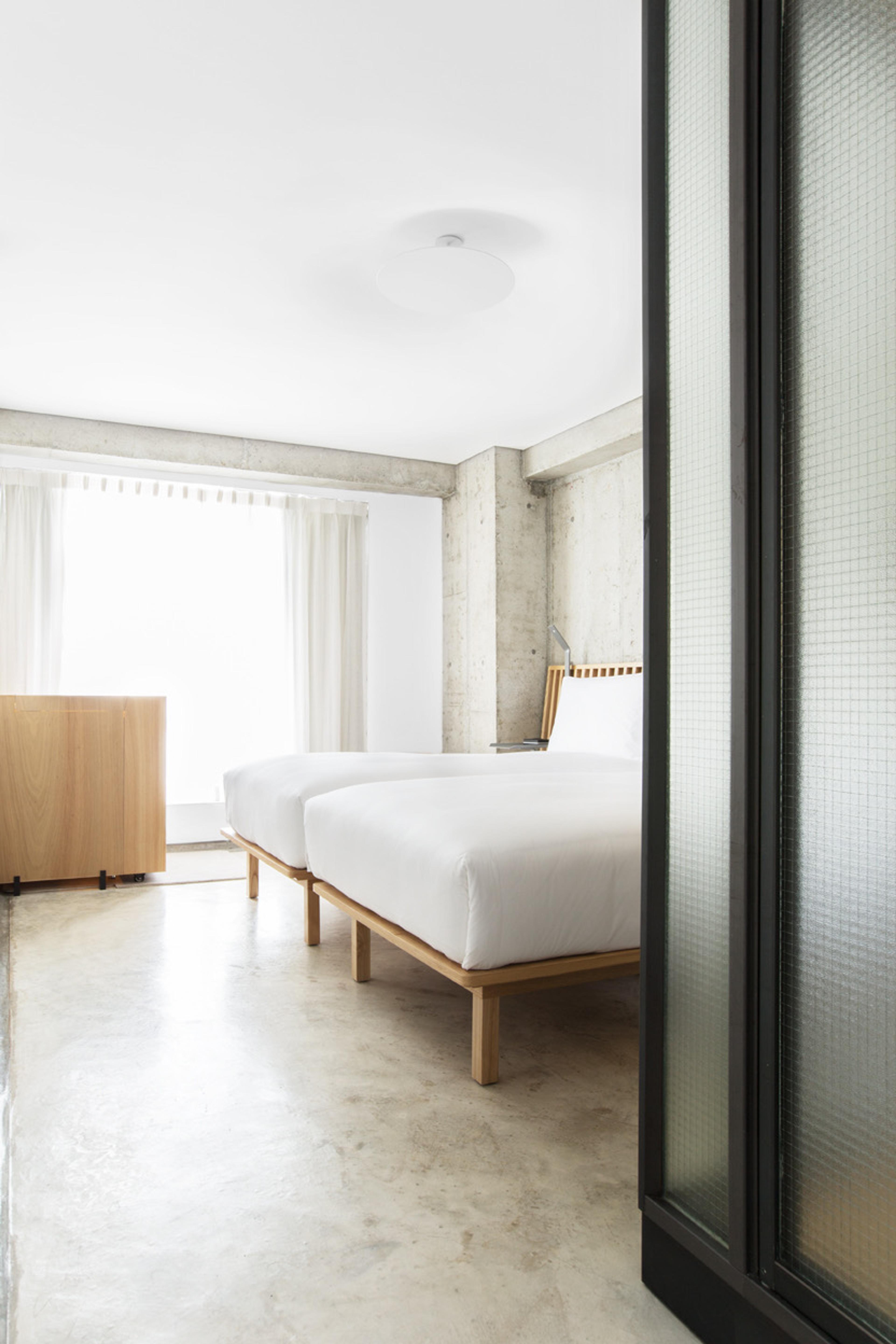 Two single beds with timber frames beneath a window with sheer curtains, beside a wooden cabinet.