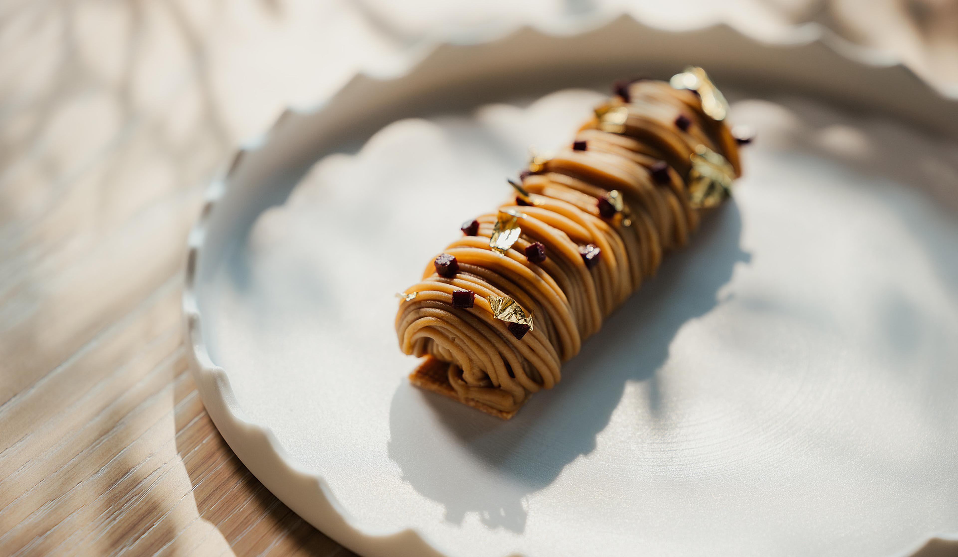 A rolled pastry dessert piped with cream and topped with gold leaf, on a white scalloped plate in dappled sunlight.