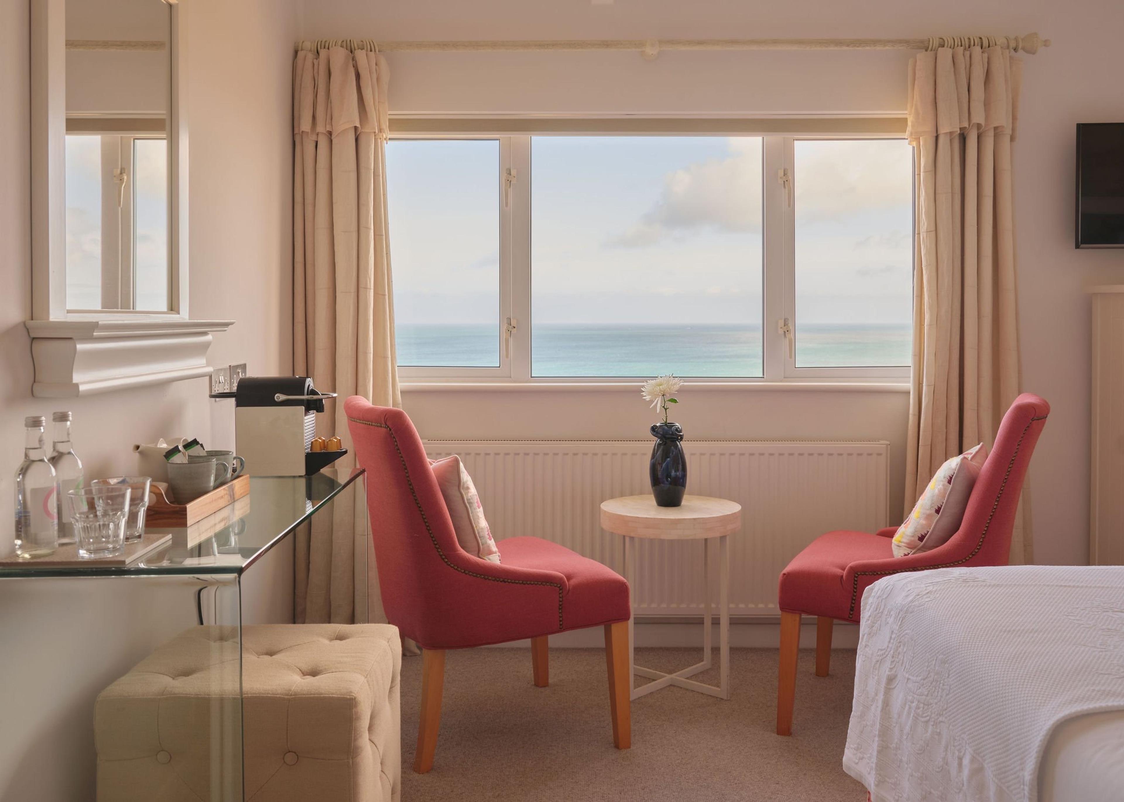 A bedroom with two red upholstered chairs around a small round table by a window filled with the sea, and the foot of a bed in the foreground.