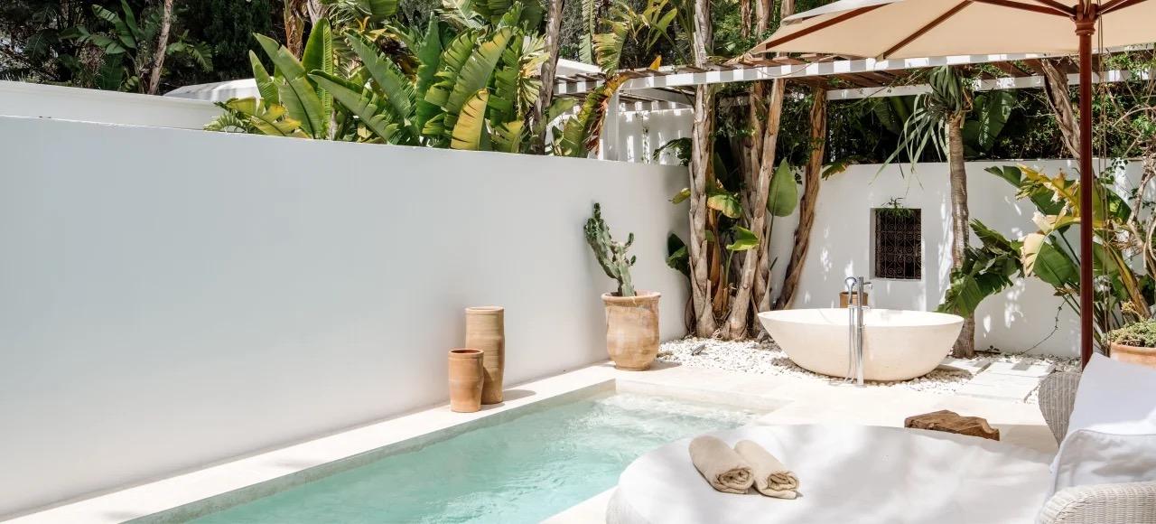 A round freestanding stone bathtub beside a small plunge pool enclosed by white walls and tropical planting, with folded towels at the edge.