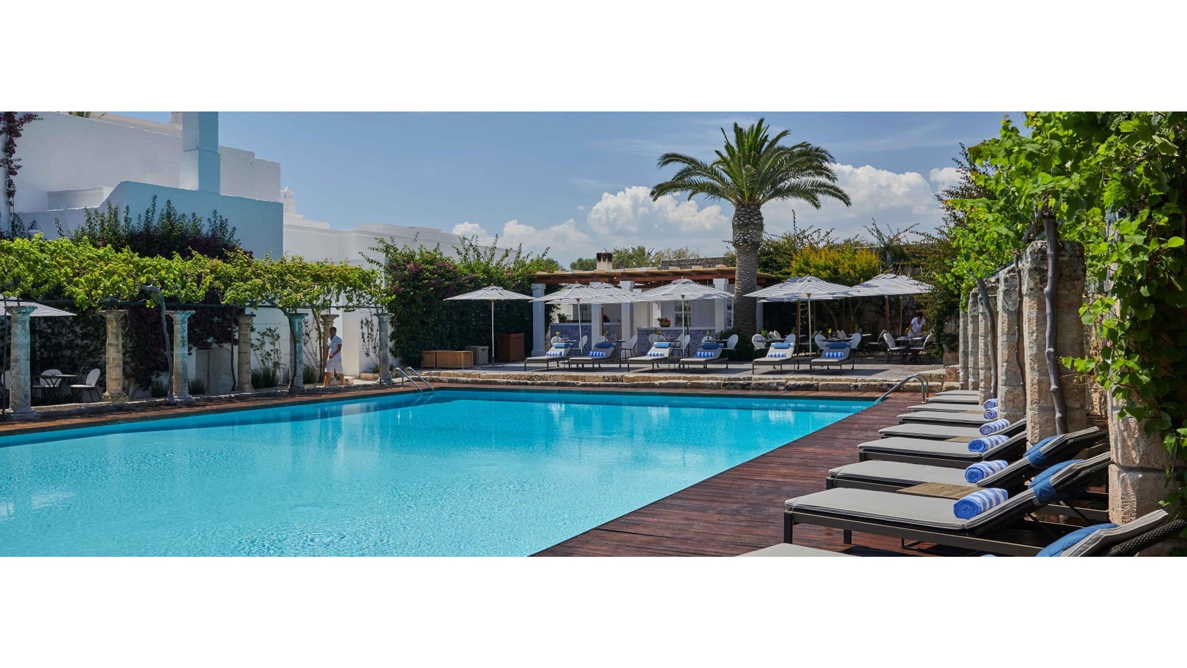 A swimming pool beside a wooden deck with sun loungers, a palm tree and a shaded bar pavilion, with one person walking poolside in the distance.