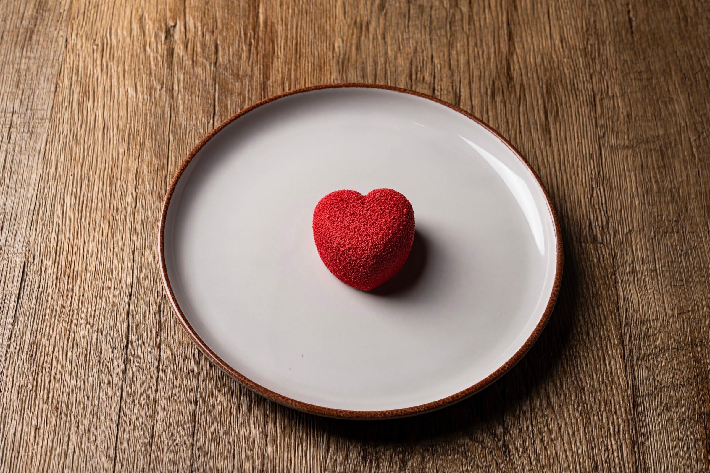 A small red heart-shaped dessert sits alone on a white plate on a wooden table.
