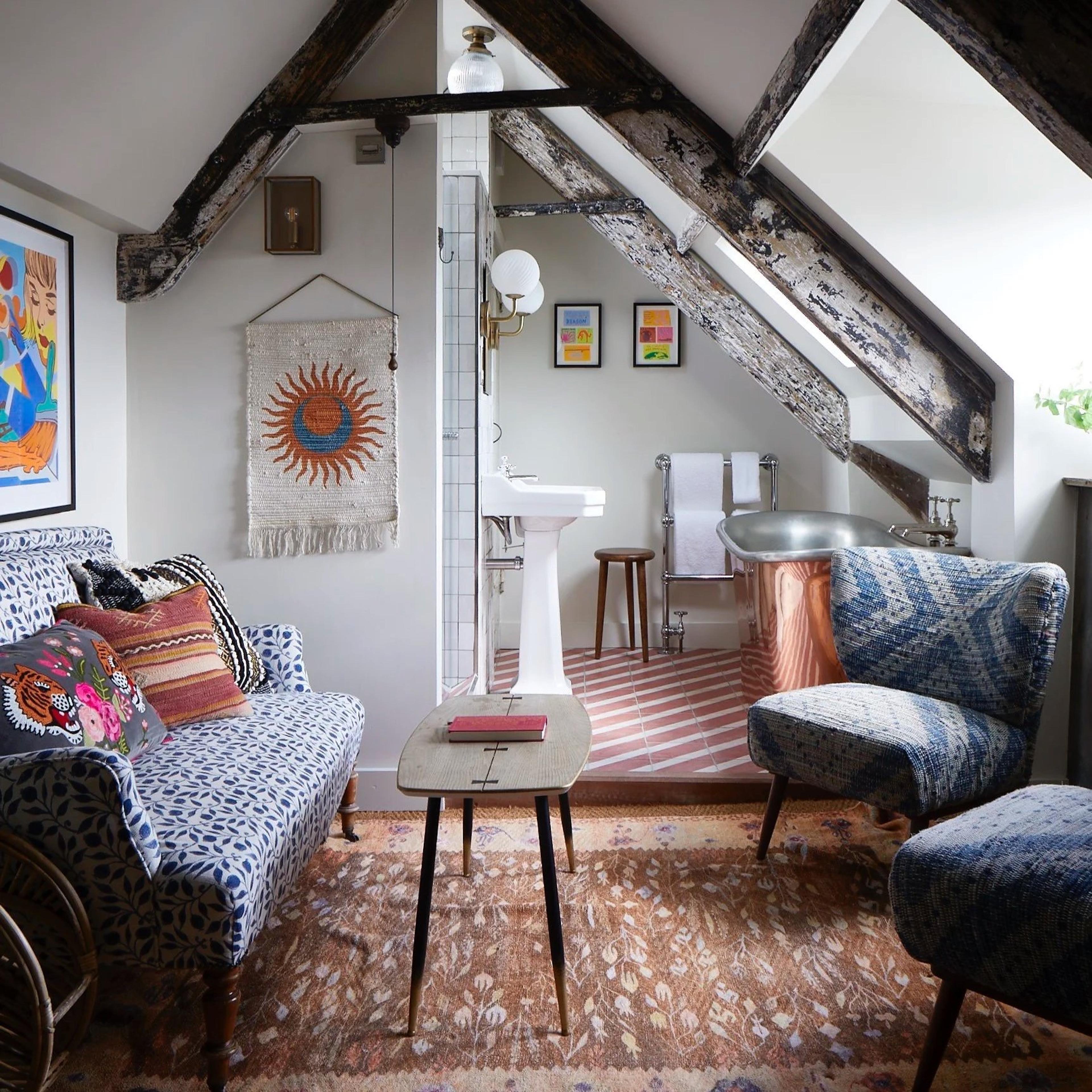 A beamed attic room with a patterned blue sofa and cushions, a small table, and a copper freestanding bath visible through an open doorway on pink and white tiled flooring.