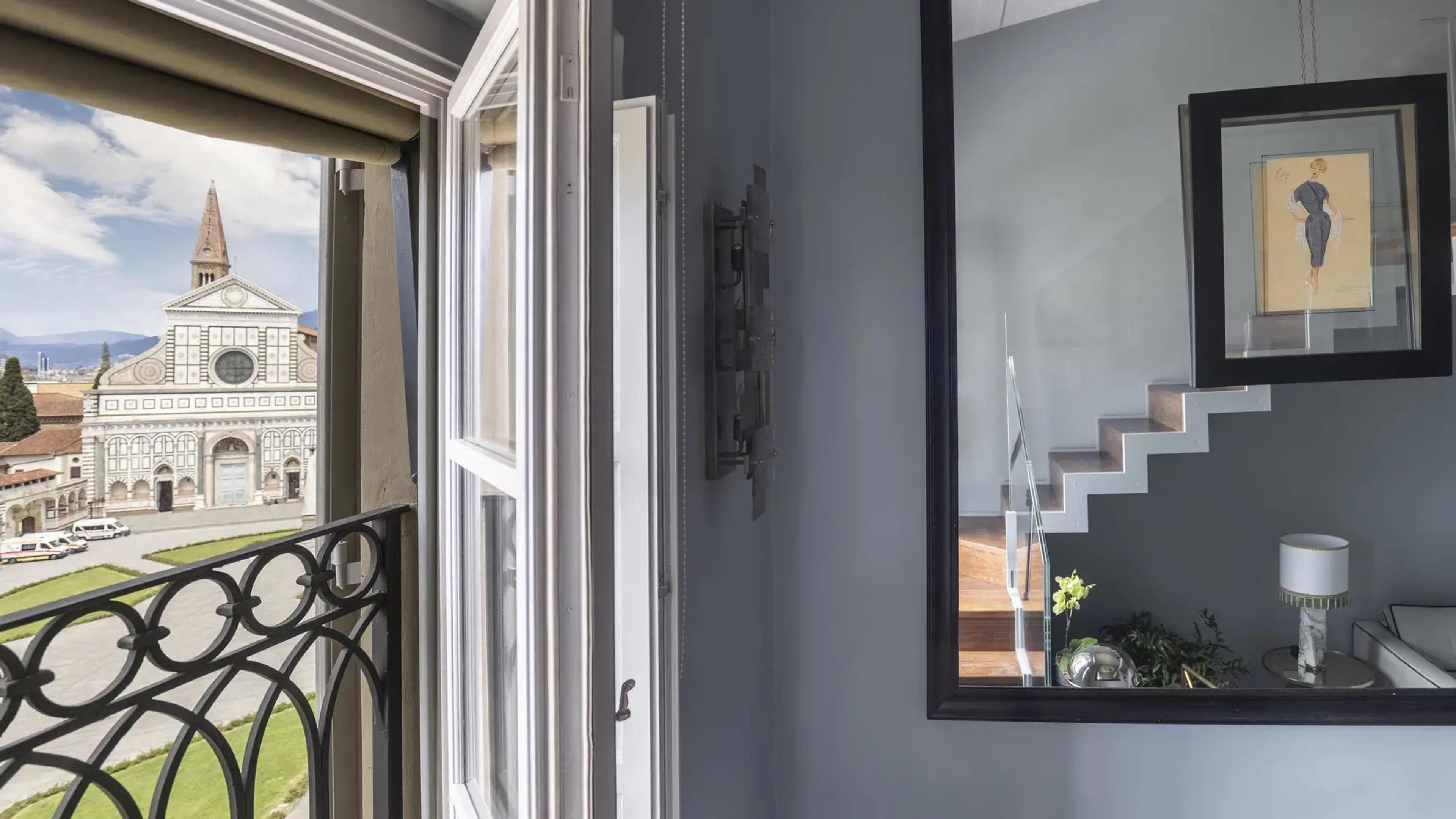 An open window with a balcony railing looking out to the facade of Santa Maria Novella, beside a mirror reflecting a staircase and a framed fashion sketch.