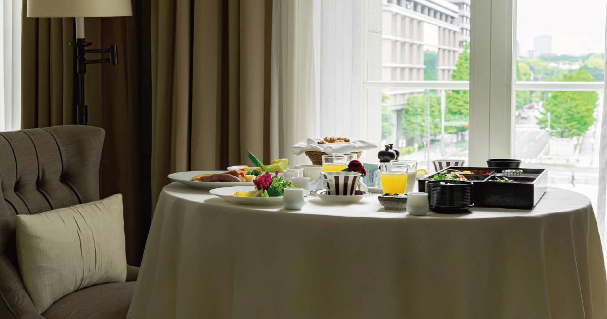 A round table laid for breakfast beside a large window overlooking trees and buildings, with a bread basket, glasses of juice and milk, and a plated dish.