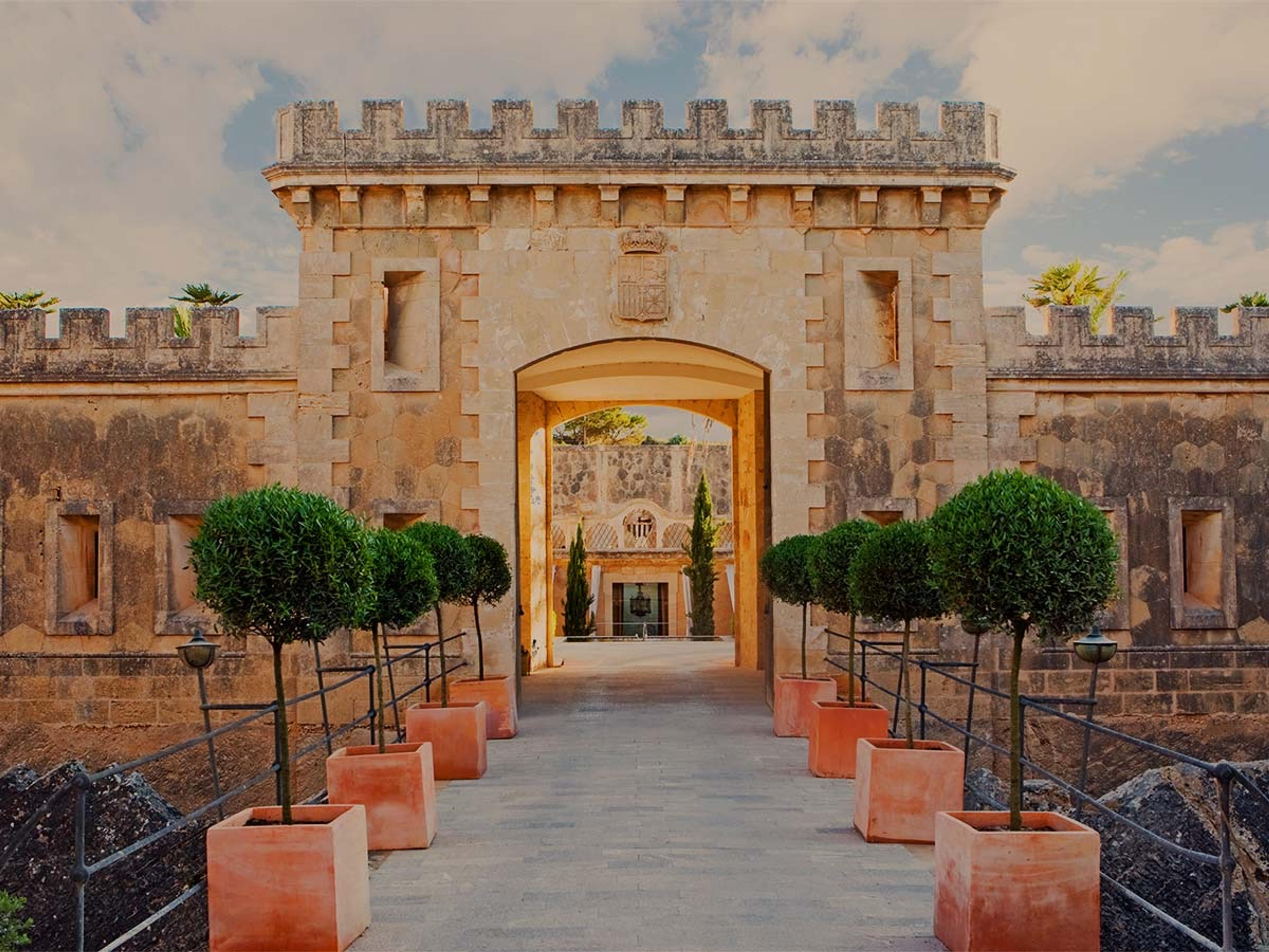 A stone fortress entrance with a crenellated tower, an arched gateway, and a row of potted topiary trees lining the path on either side.
