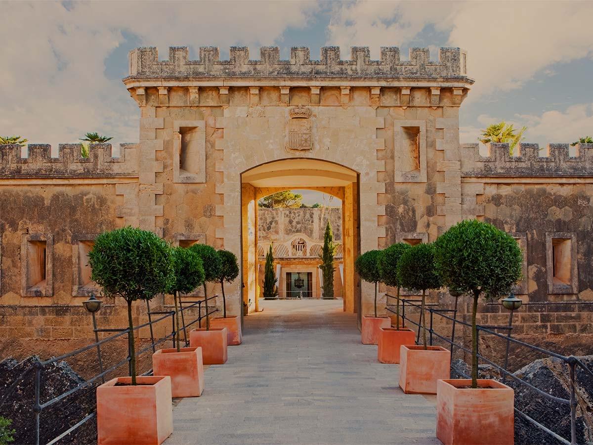 A stone fortress entrance with a crenellated tower, an arched gateway, and a row of potted topiary trees lining the path on either side.
