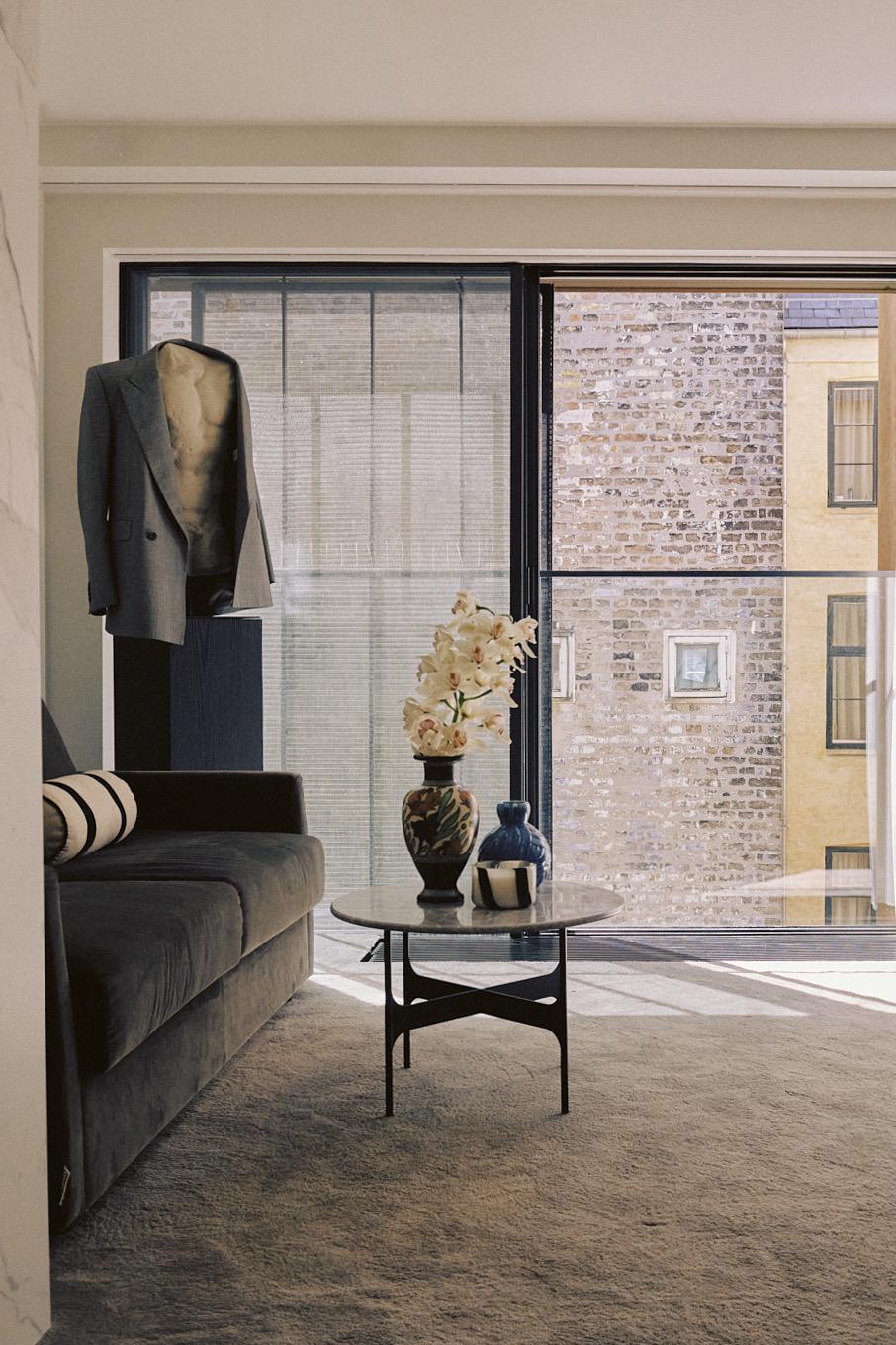 A living room with a dark sofa, a blazer hung over a speaker, and a side table holding a vase of flowers, in front of a glass door looking onto a brick building across the street.