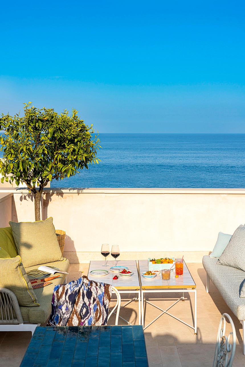 A rooftop terrace with two sofas around a low table set with wine, fruit and drinks, a potted tree, and the sea meeting the sky in the distance.