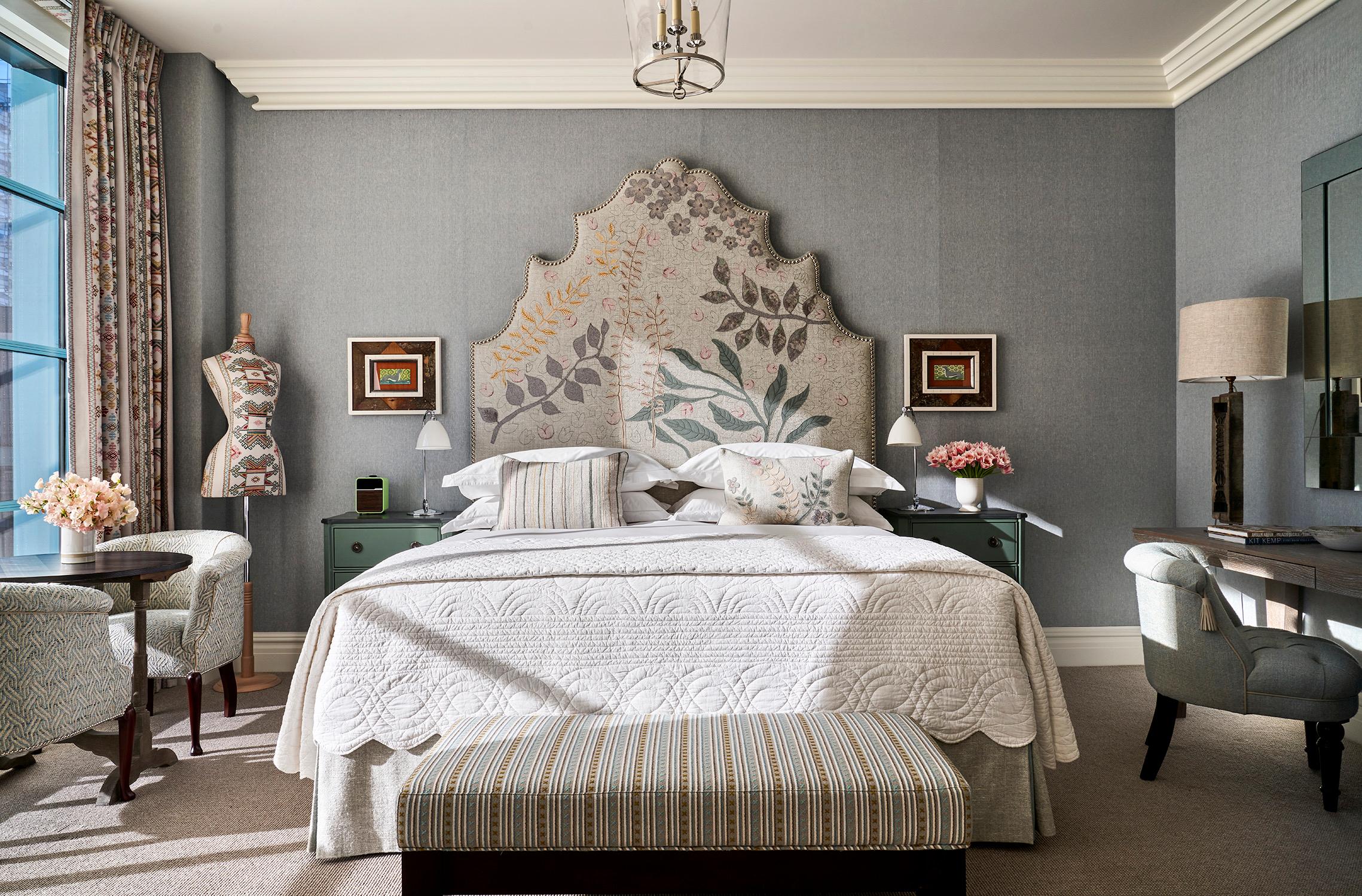 A bedroom with a tall floral upholstered headboard, a dressmaker's mannequin beside the window, and a crystal chandelier overhead.