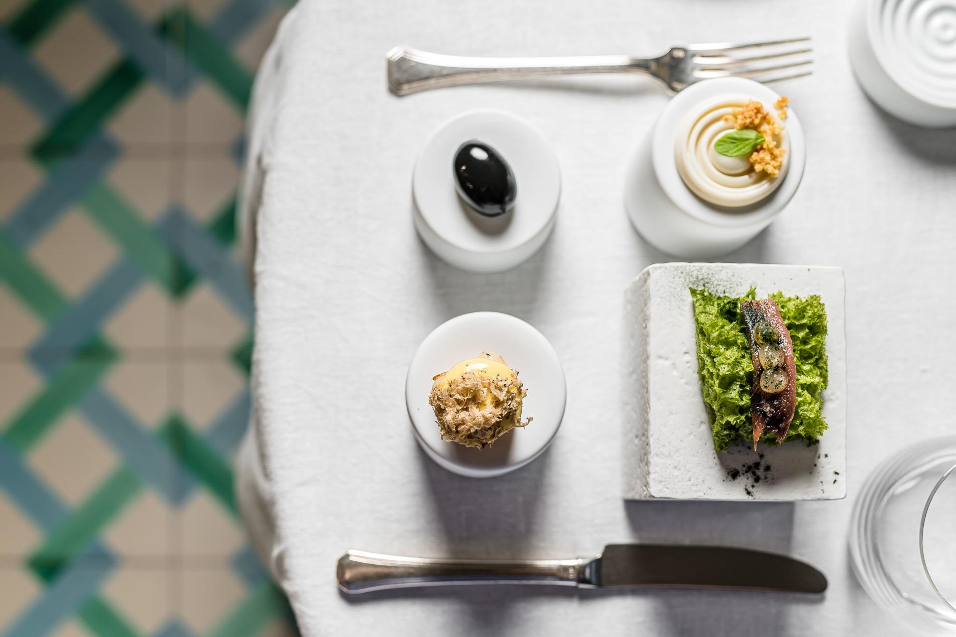 Small plates of food laid on a white tablecloth, including a dish of greens topped with fish, with a fork and knife beside them and a trellis blurred behind.
