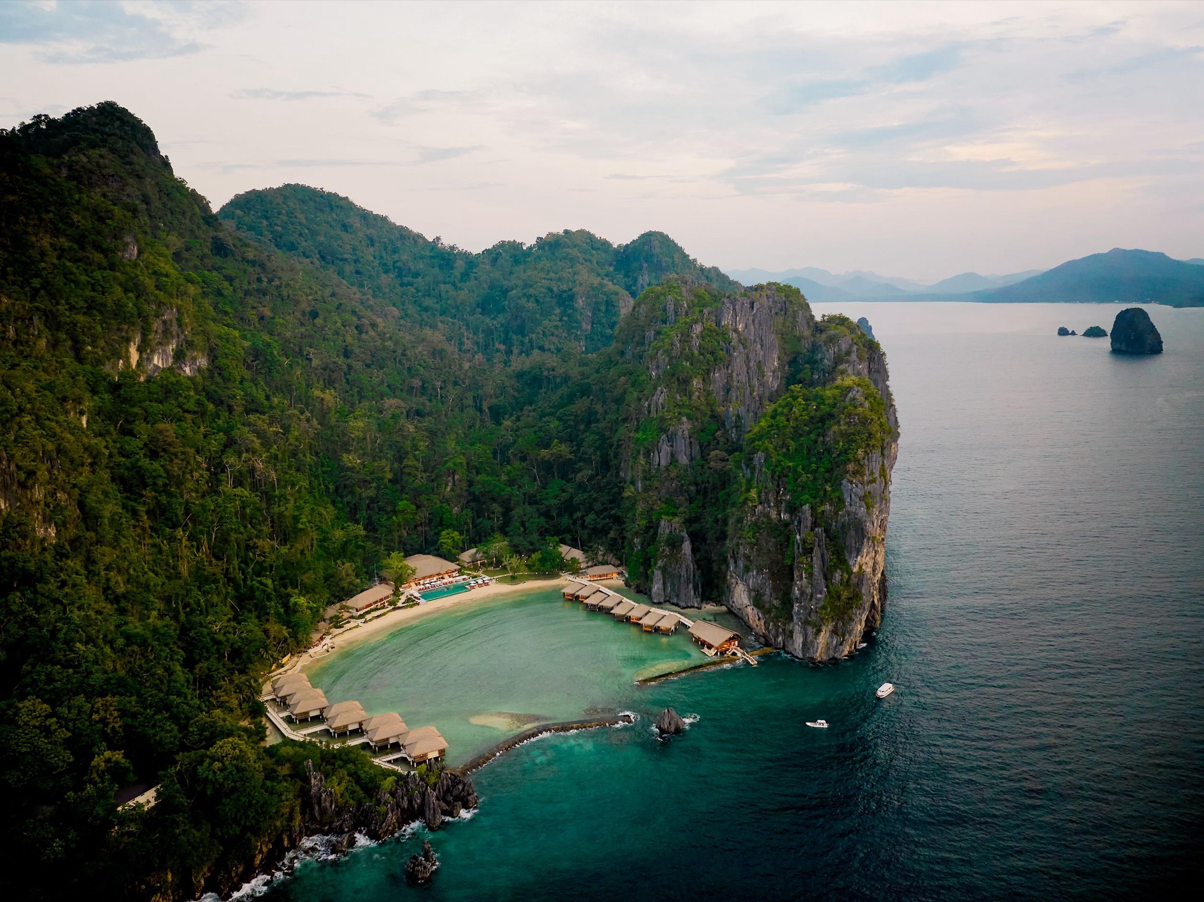 An aerial view of a forested limestone island, a pale lagoon held in its cove and the open sea beyond.