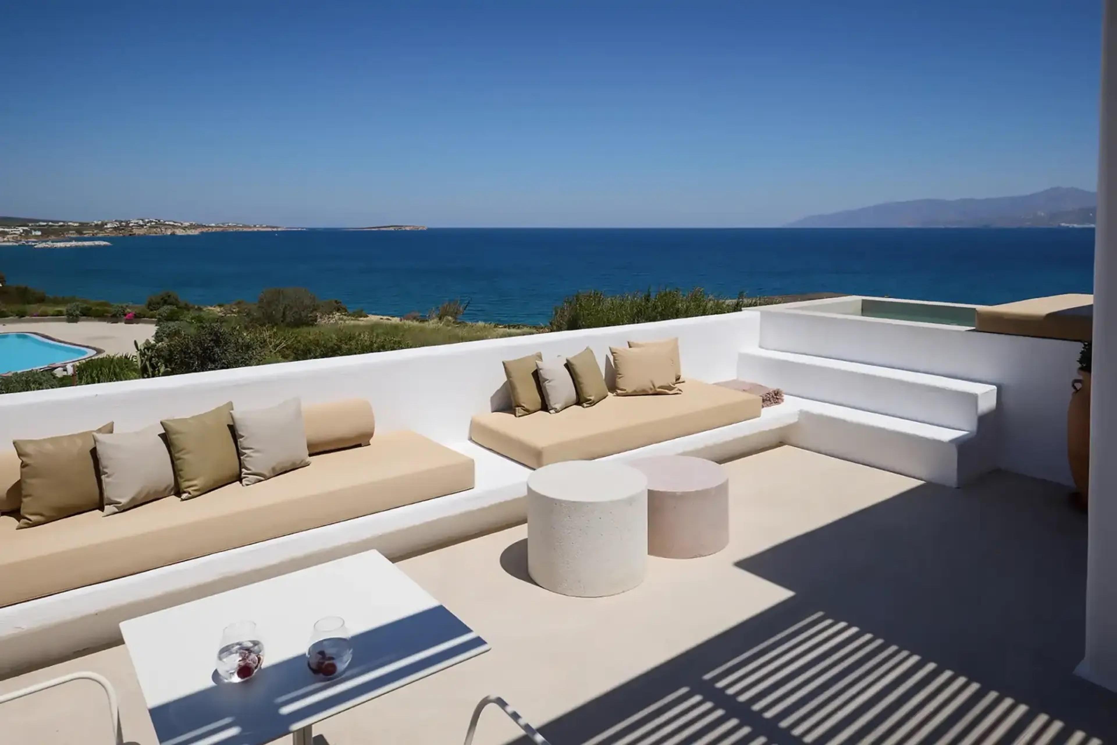 A covered terrace lounge with cushioned bench seating and a low table set with two drinks, looking out over a pool and the sea beyond.