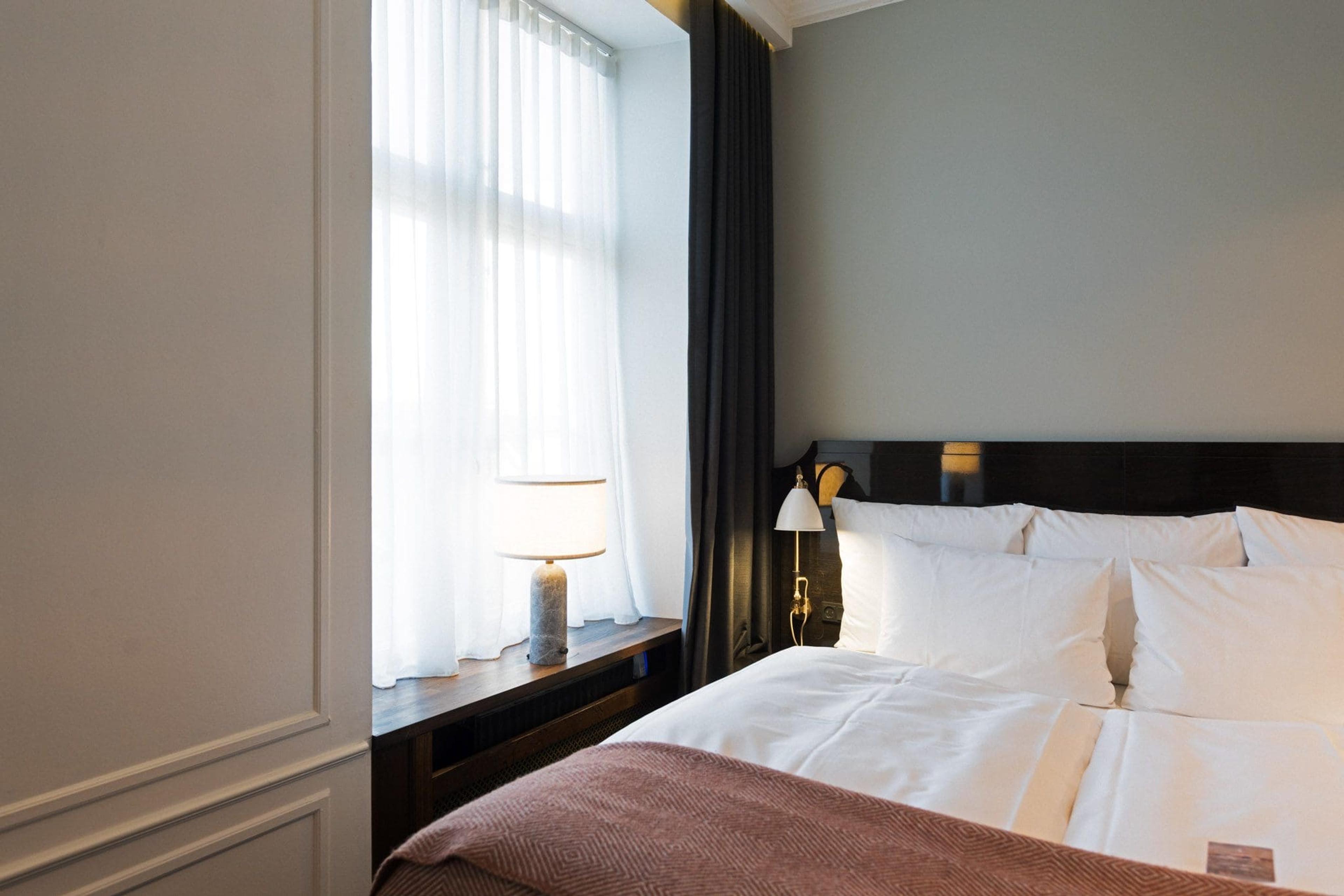 A bed with white pillows and a rose-coloured throw beside a curtained window with a lit table lamp.