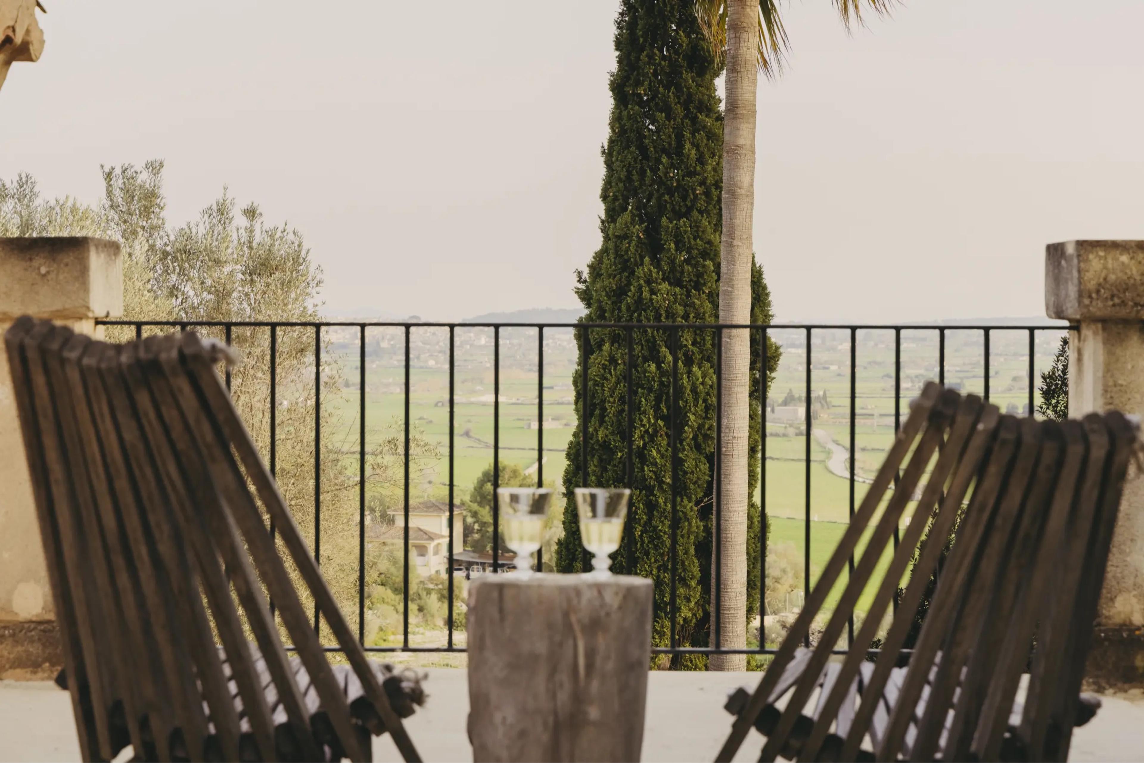 Two empty wooden deck chairs and two glasses of wine on a tree-stump table, looking out over a railing to fields and hills.