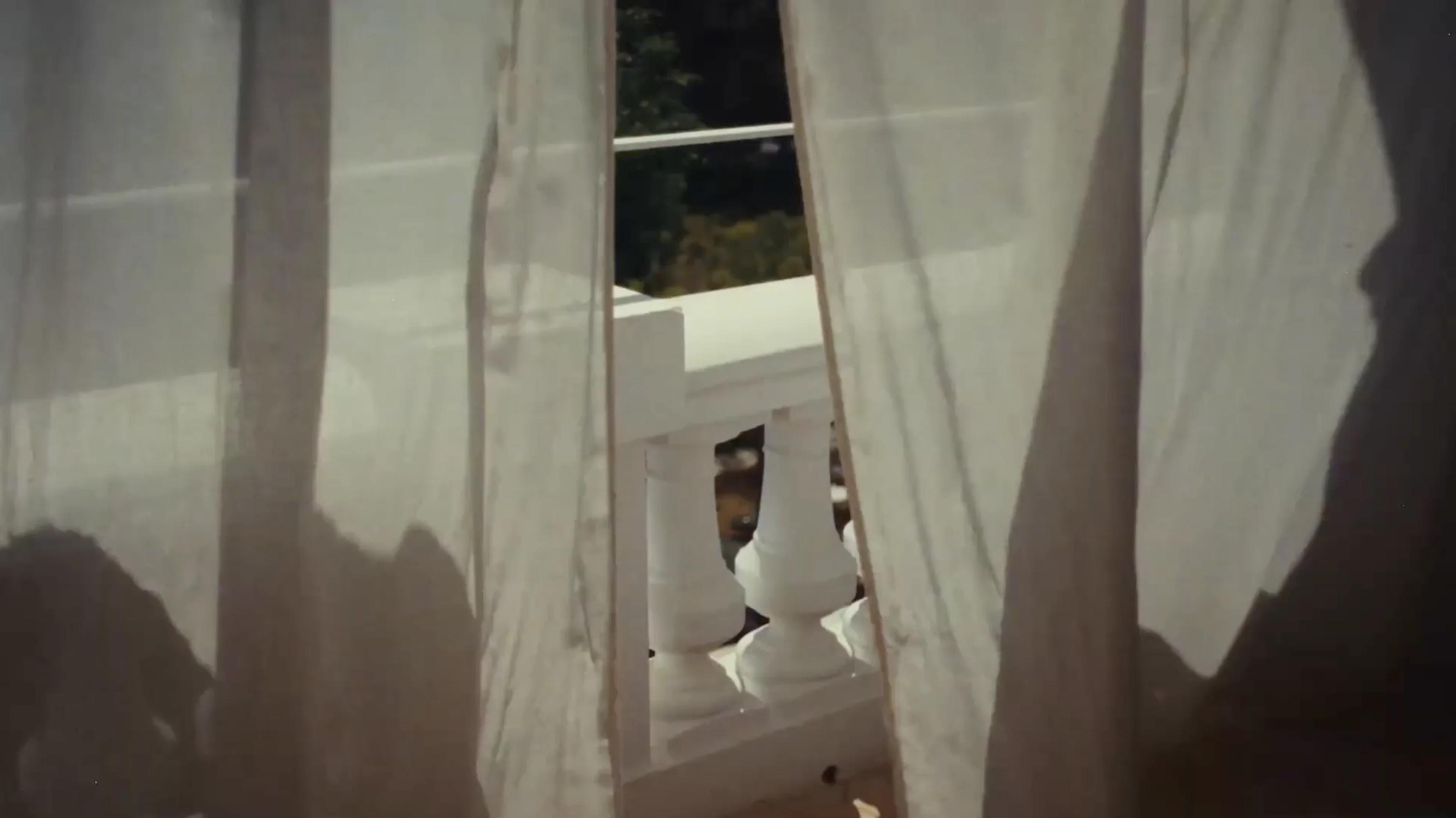 Sheer white curtains blowing in front of an open doorway, with white balcony balusters and greenery visible beyond.