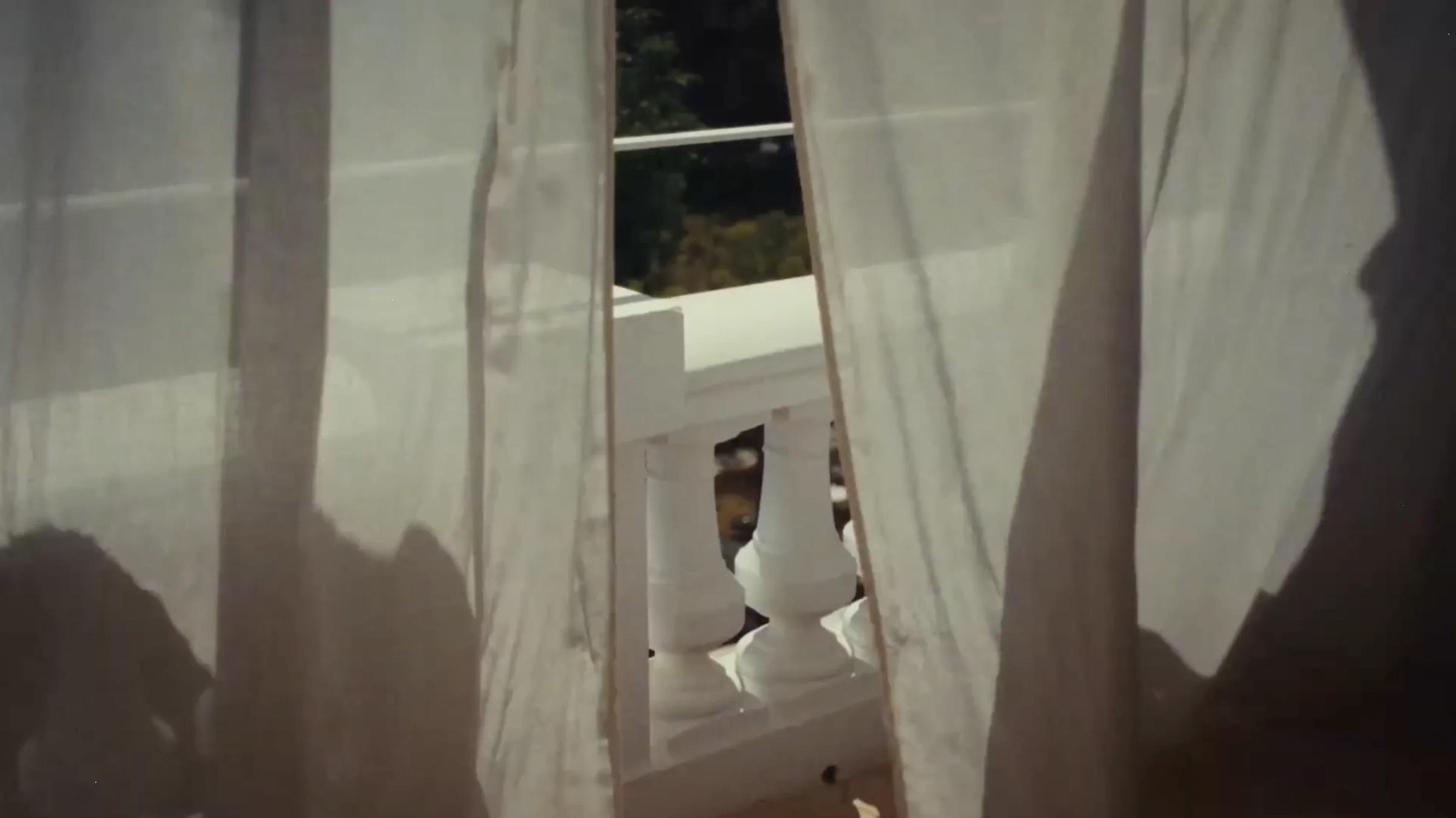 Sheer white curtains blowing in front of an open doorway, with white balcony balusters and greenery visible beyond.