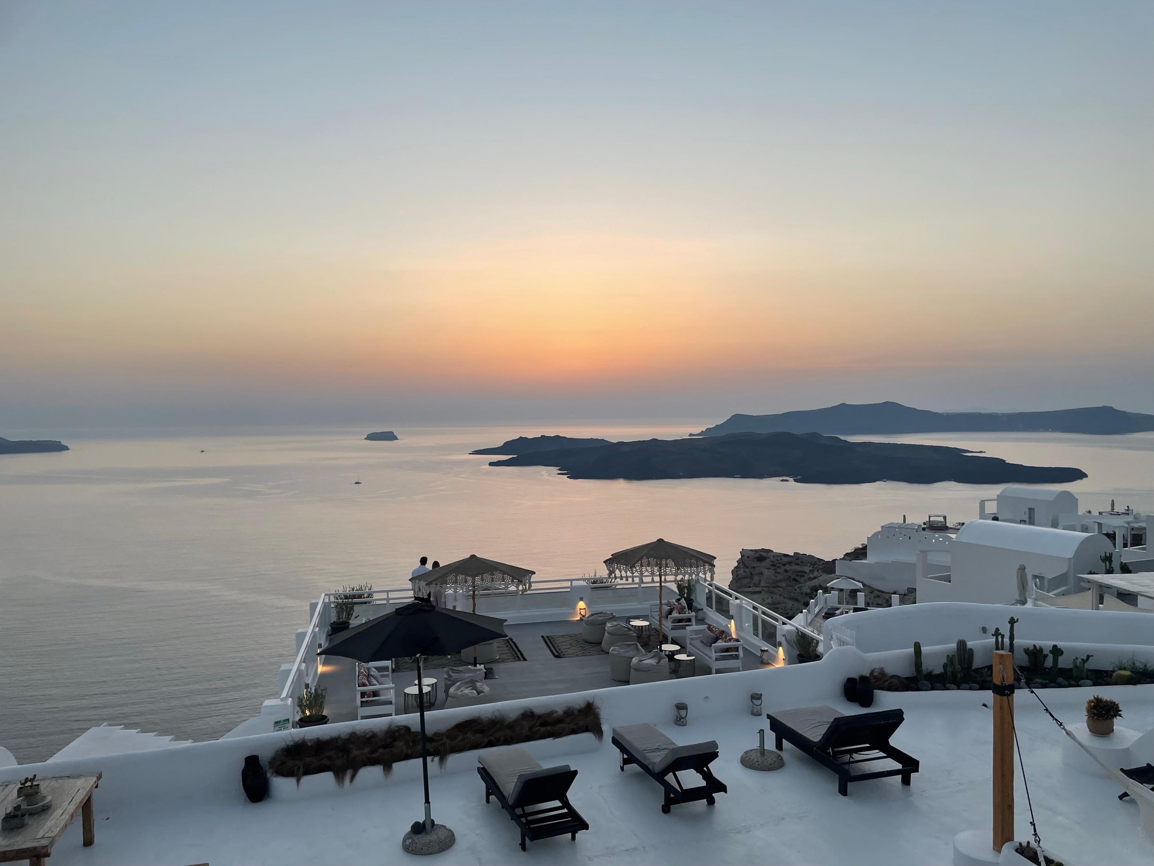 Sunset over the Santorini caldera, islands dark on the water beyond white rooftop terraces and sun loungers