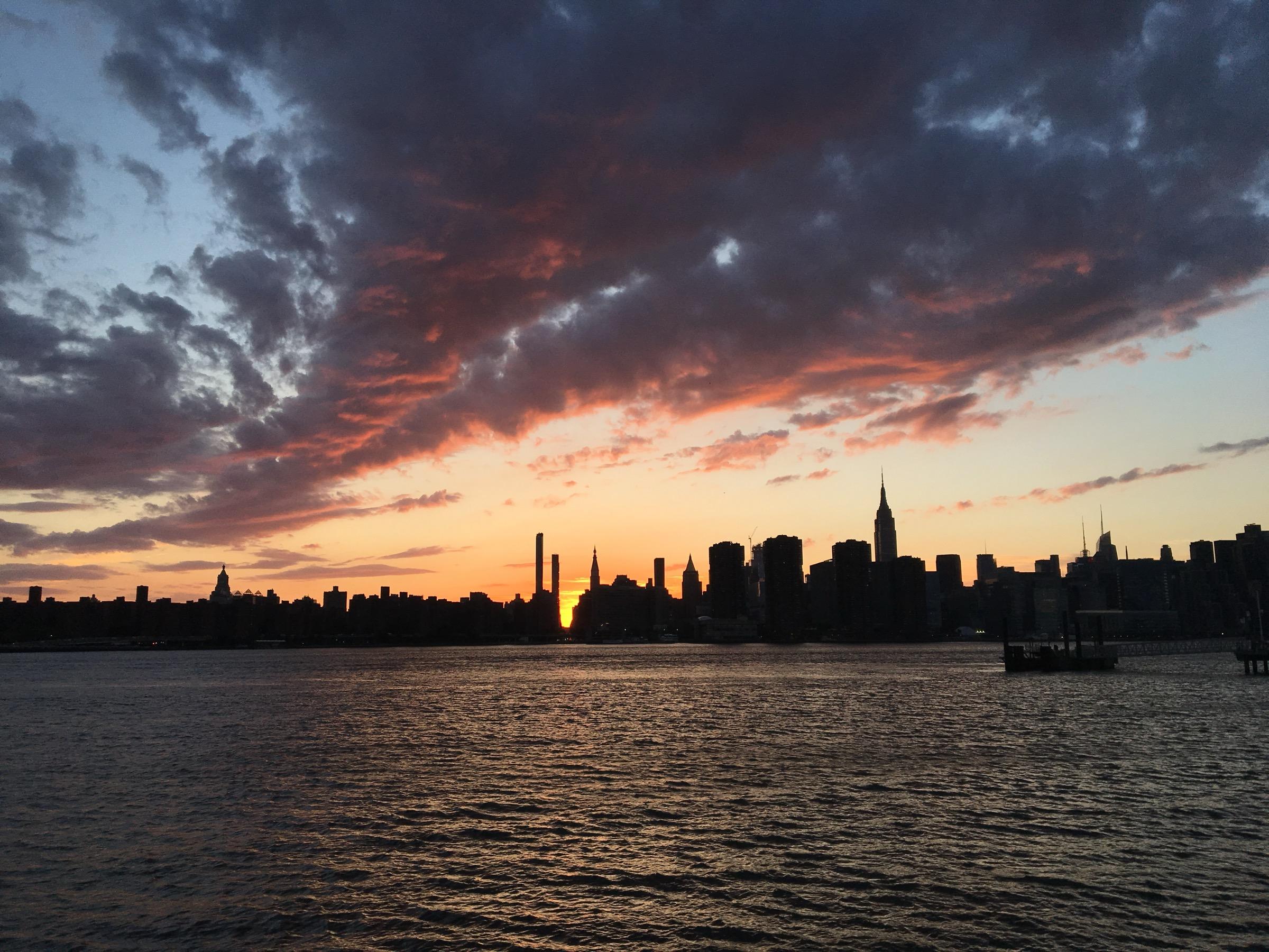 The Midtown Manhattan skyline in silhouette at sunset across the East River from Brooklyn, the Empire State Building against red-lit clouds