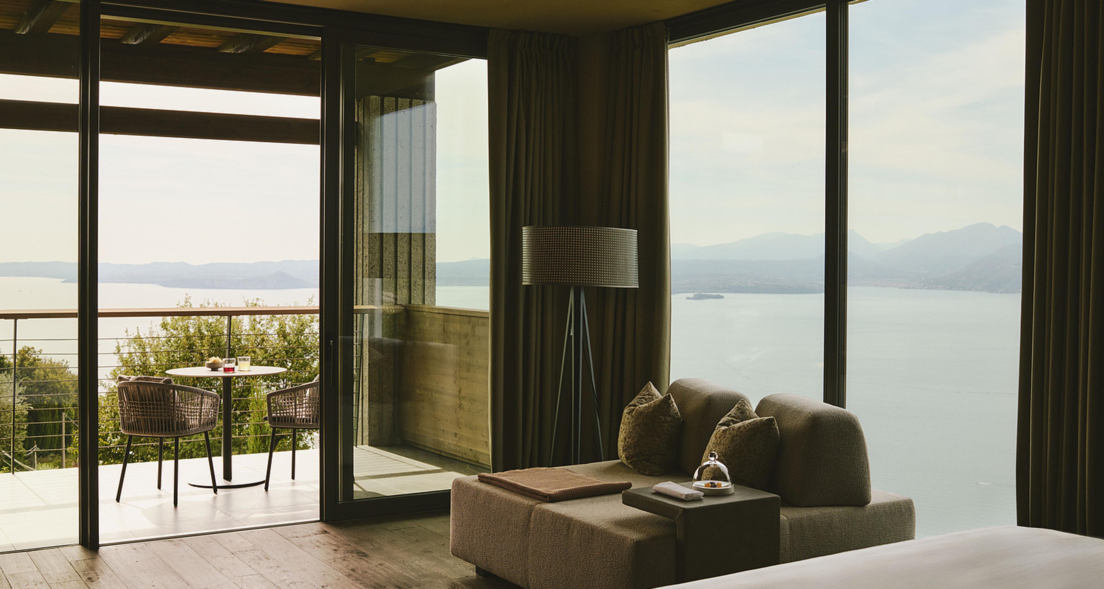 A hotel room with a daybed and lamp beside floor to ceiling windows looking over Lake Garda, a balcony table outside.