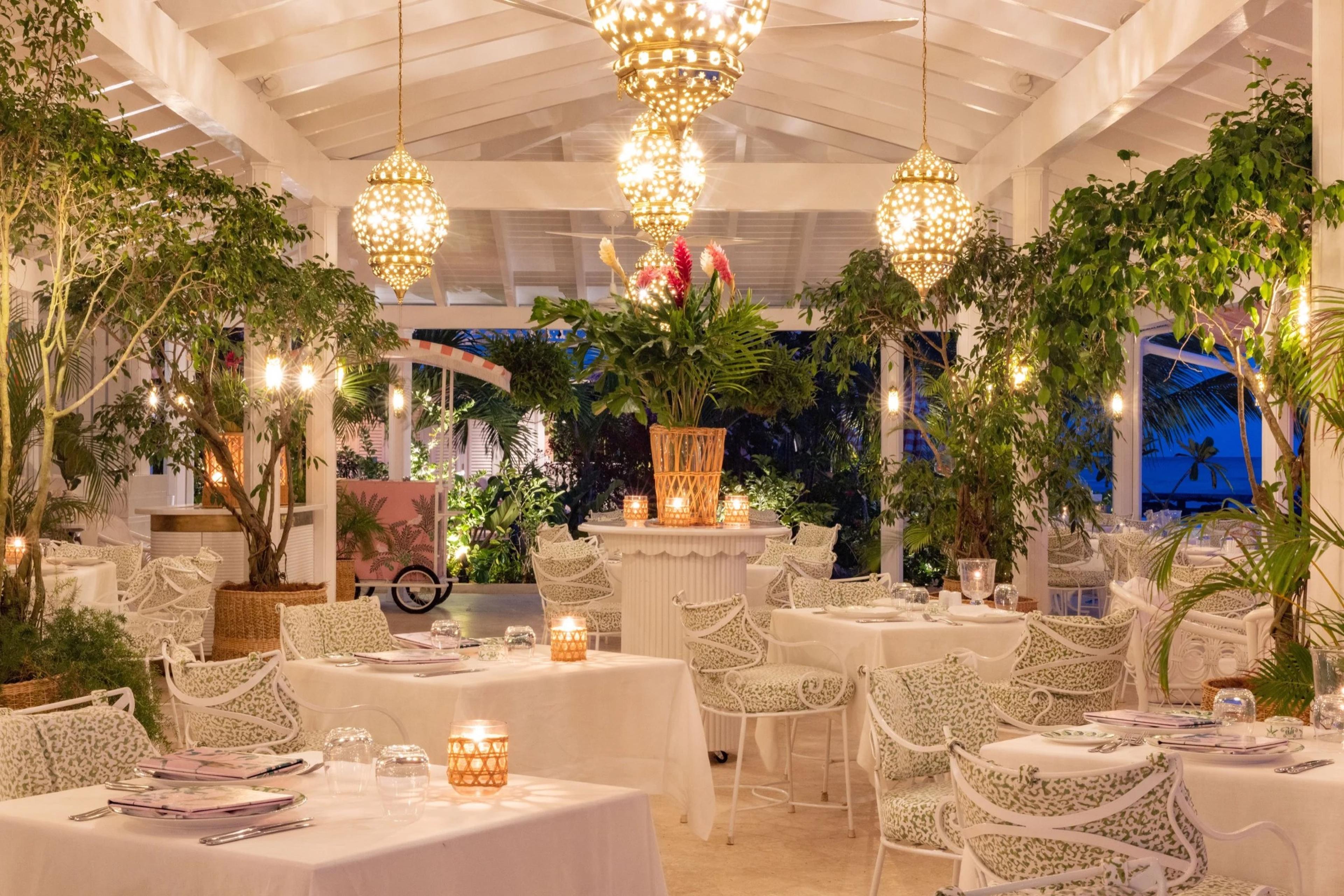 an outdoor restaurant pavilion at night, tables laid in white linen under hanging lanterns and trees strung with lights