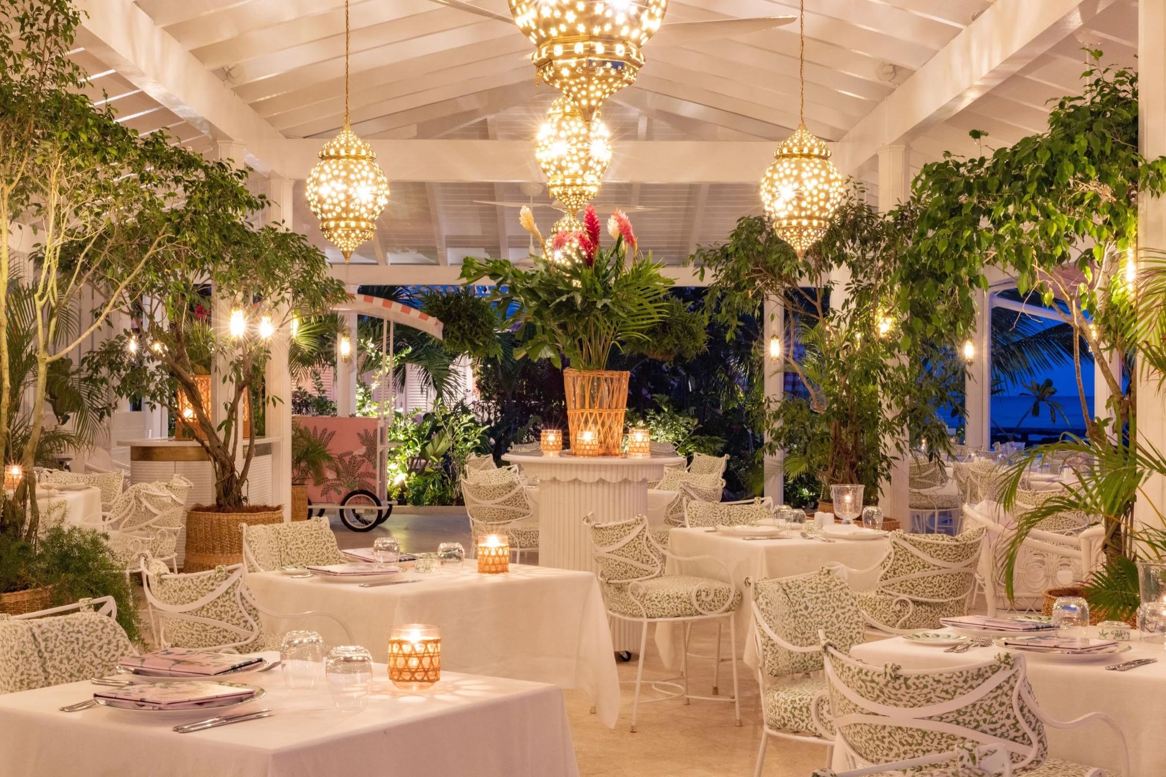 an outdoor restaurant pavilion at night, tables laid in white linen under hanging lanterns and trees strung with lights