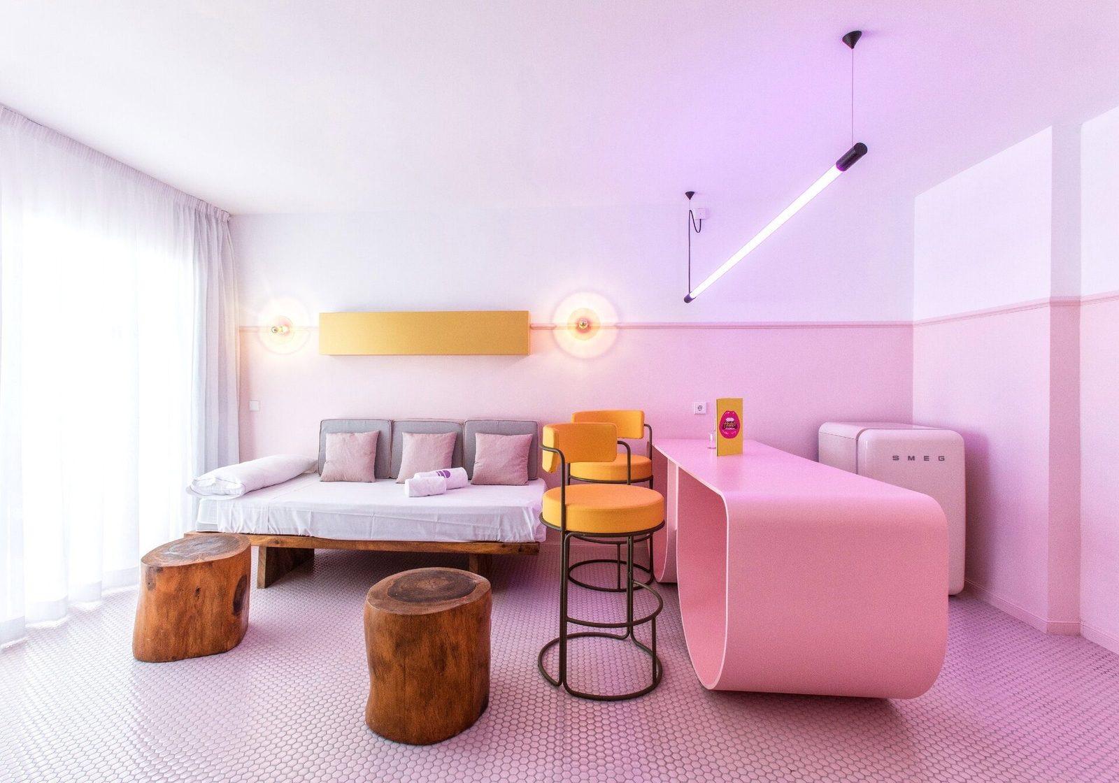 A pink room with a low sofa bed on a wooden base, two tree-trunk stools, a yellow-seated bar stool, a curved pink desk, a pink retro fridge and a hanging linear light, on a floor of small pink tiles.